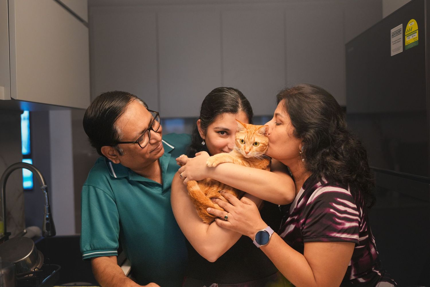 Family of three sharing a loving moment with an orange tabby cat in a modern kitchen.