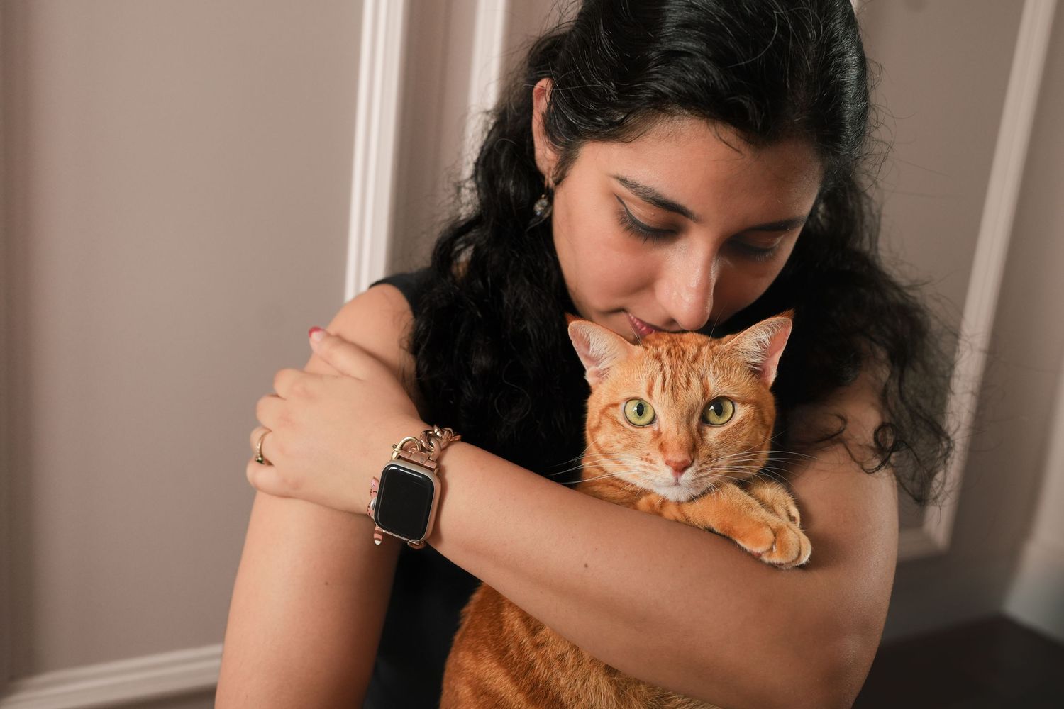 Young woman with dark hair and smartwatch lovingly hugging an orange tabby cat indoors.
