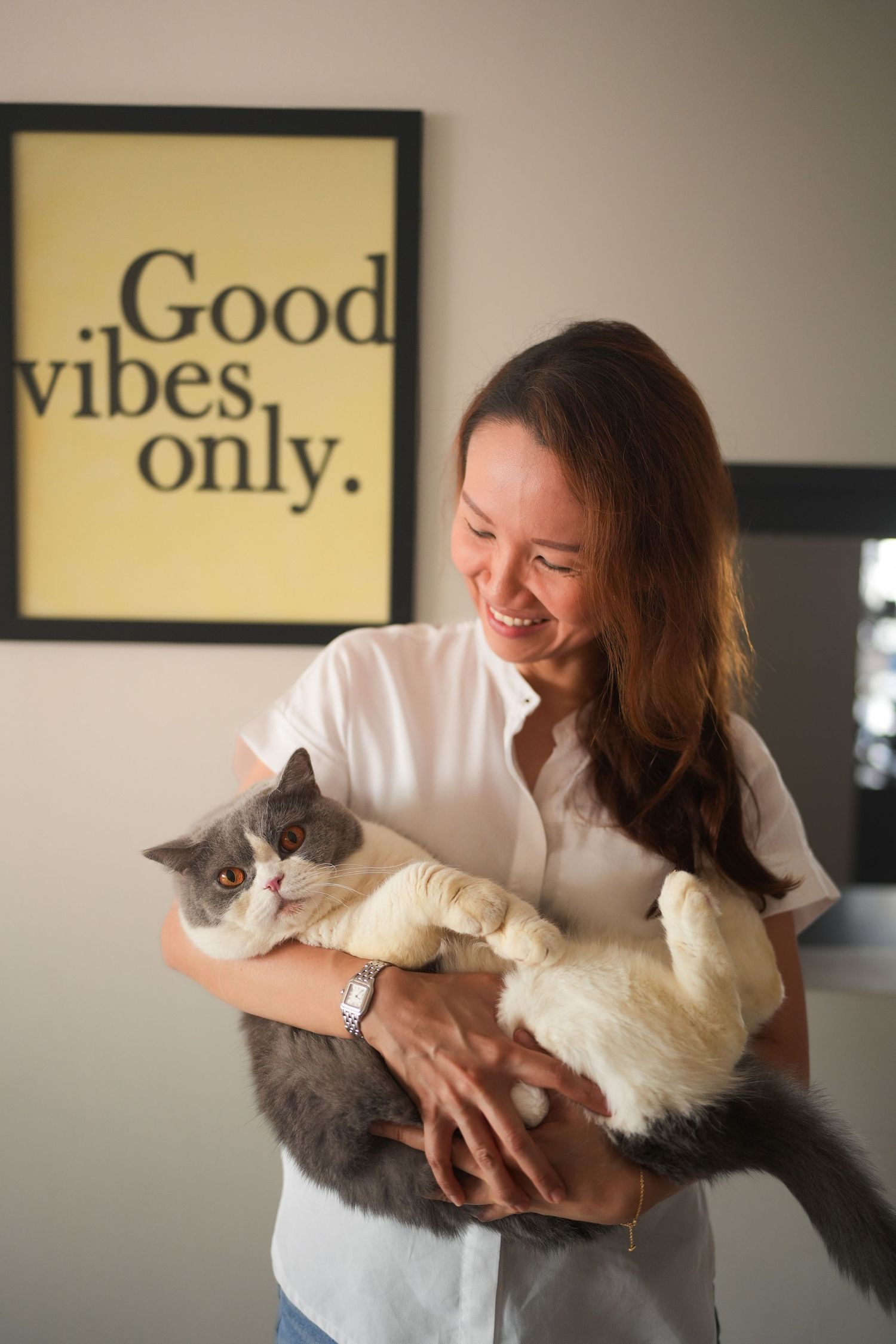 Smiling Asian woman holding a fluffy gray and white cat beneath a Good Vibes Only wall sign.