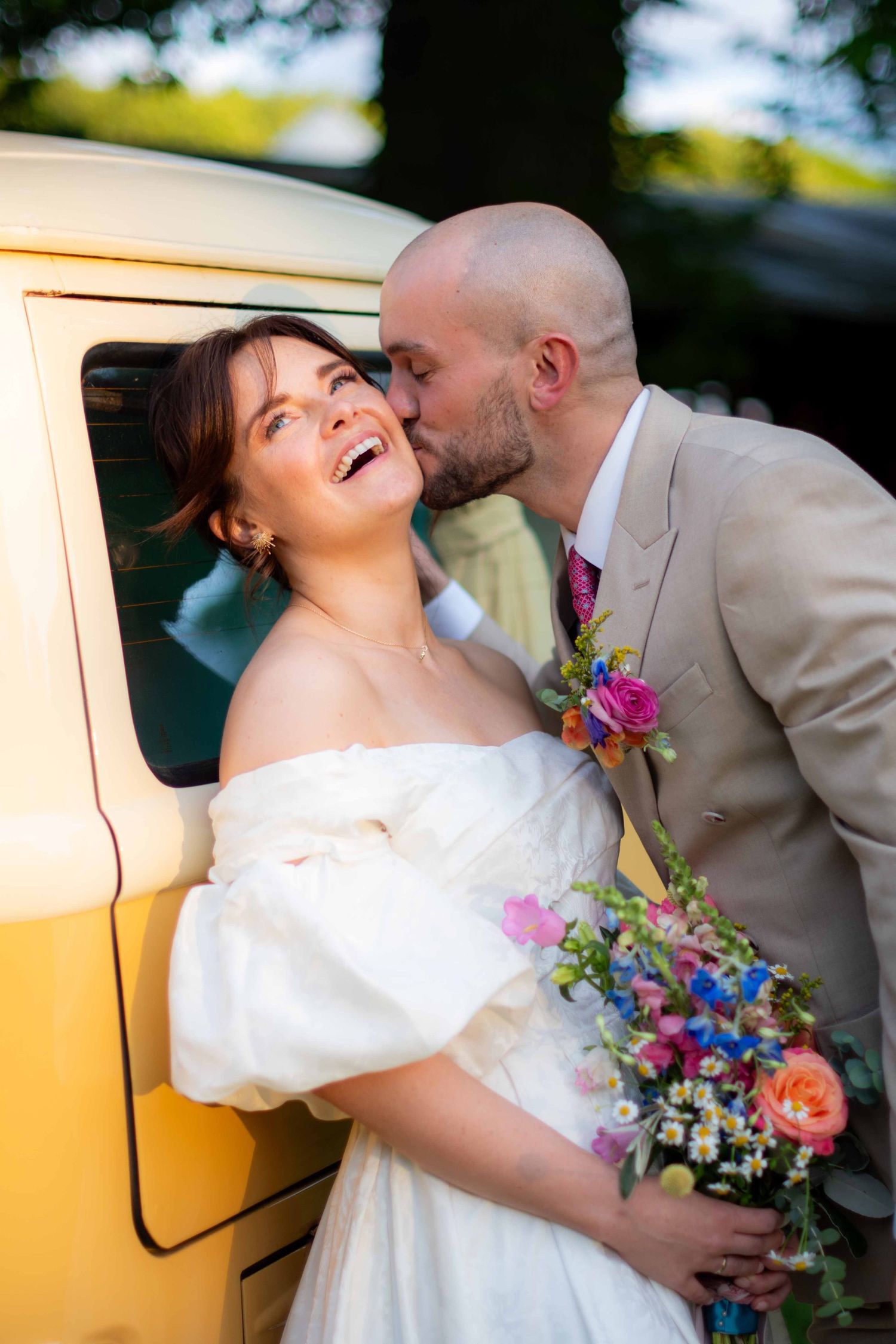 Portrait photo of the wedding groom kissing his bride, it is sharp with high contrast and a blurry background. The bride is laughing.