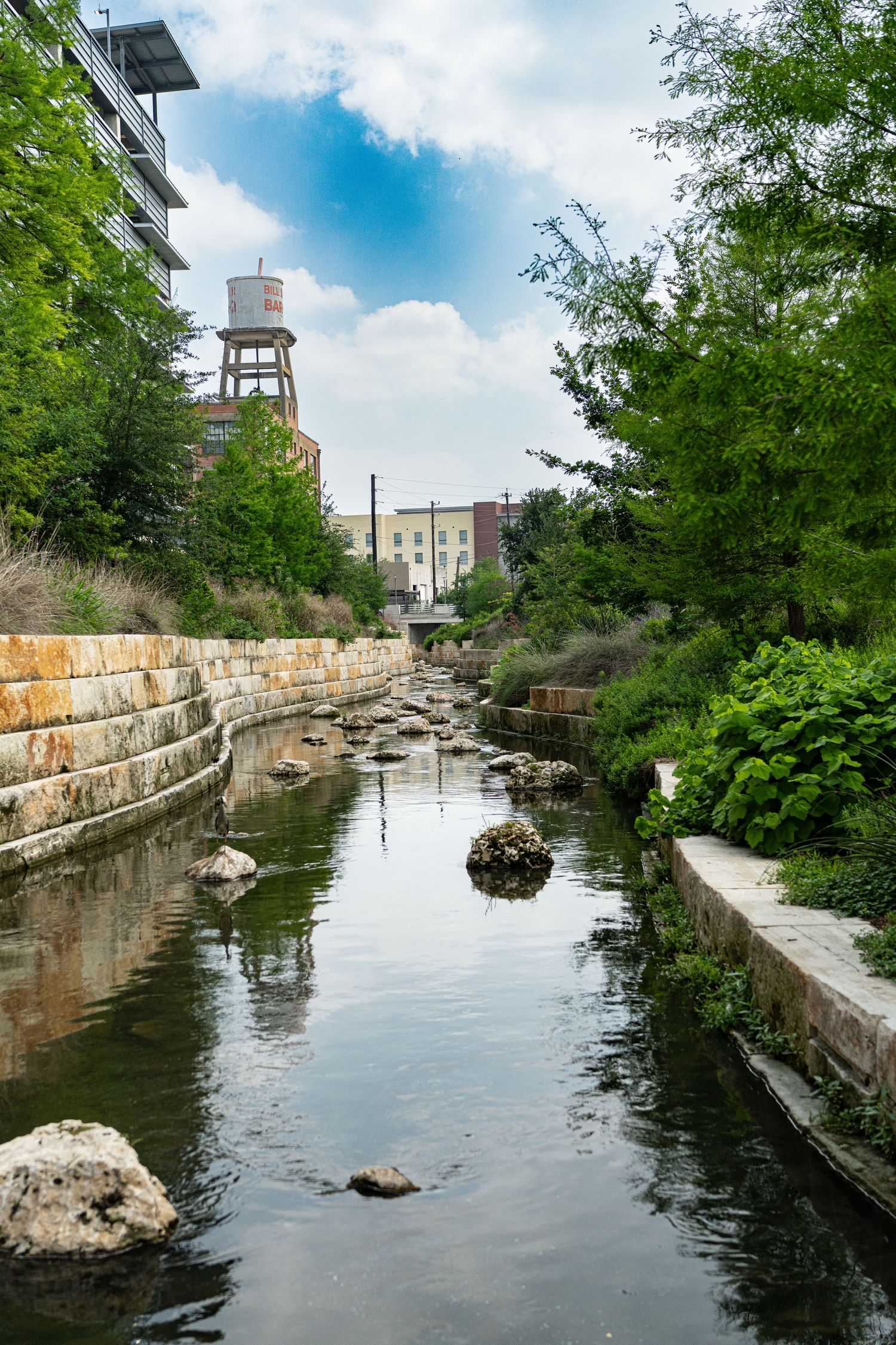 Urban creek with stone retaining walls, lush trees, and a historic water tower in the background under a partly cloudy sky.