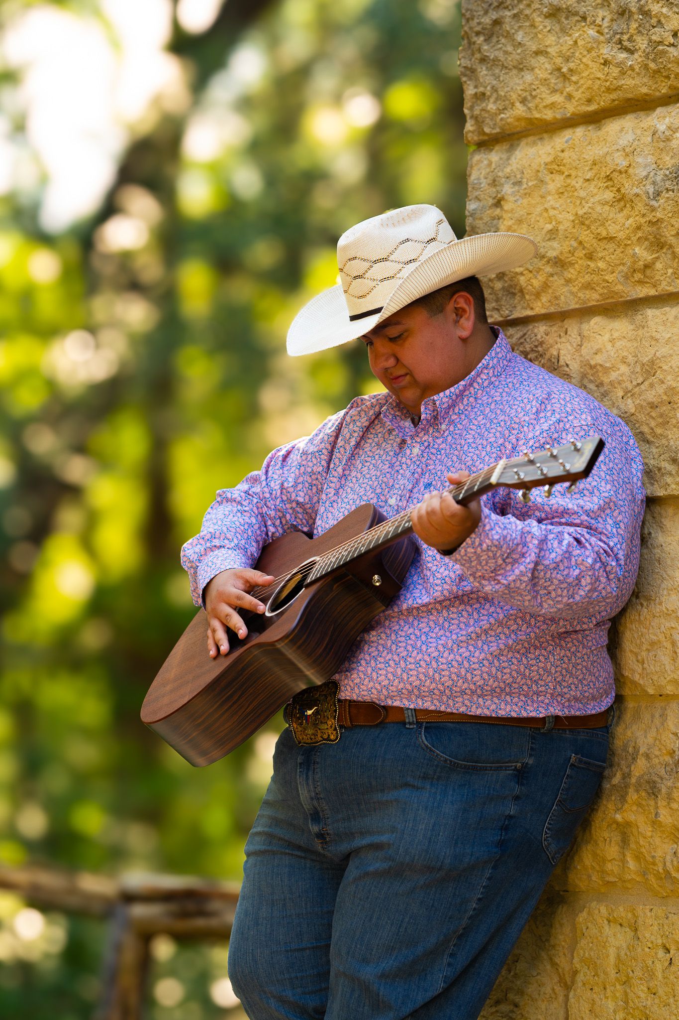 Man in cowboy hat and floral shirt playing acoustic guitar while leaning against stone wall outdoors.