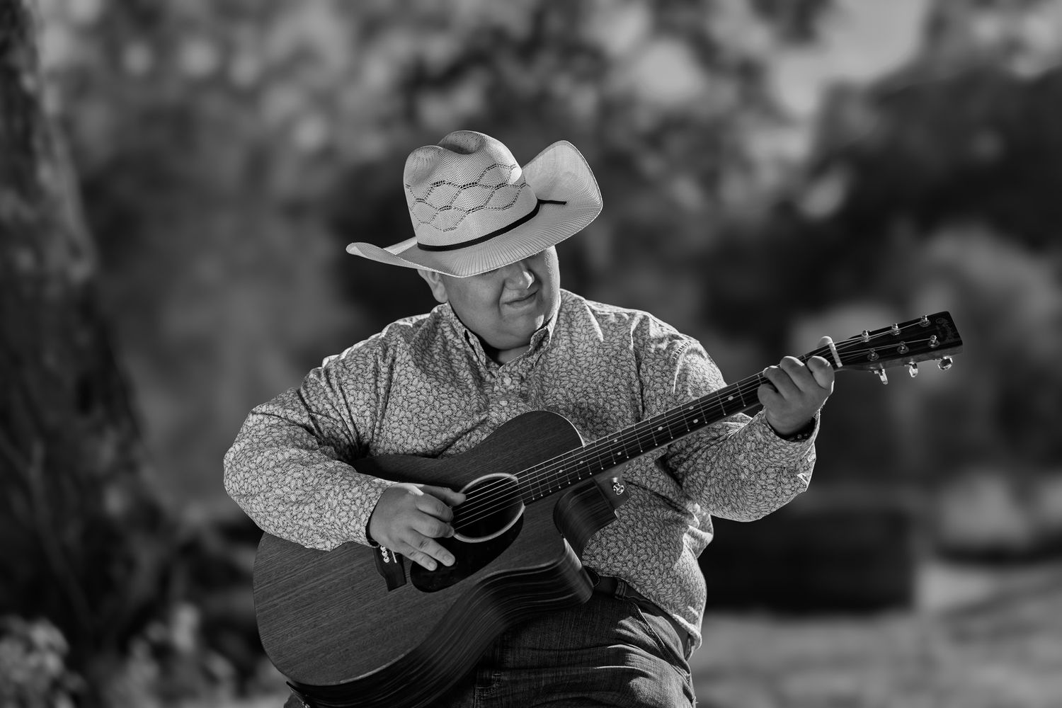 Black and white photo of a man in a cowboy hat playing acoustic guitar outdoors.