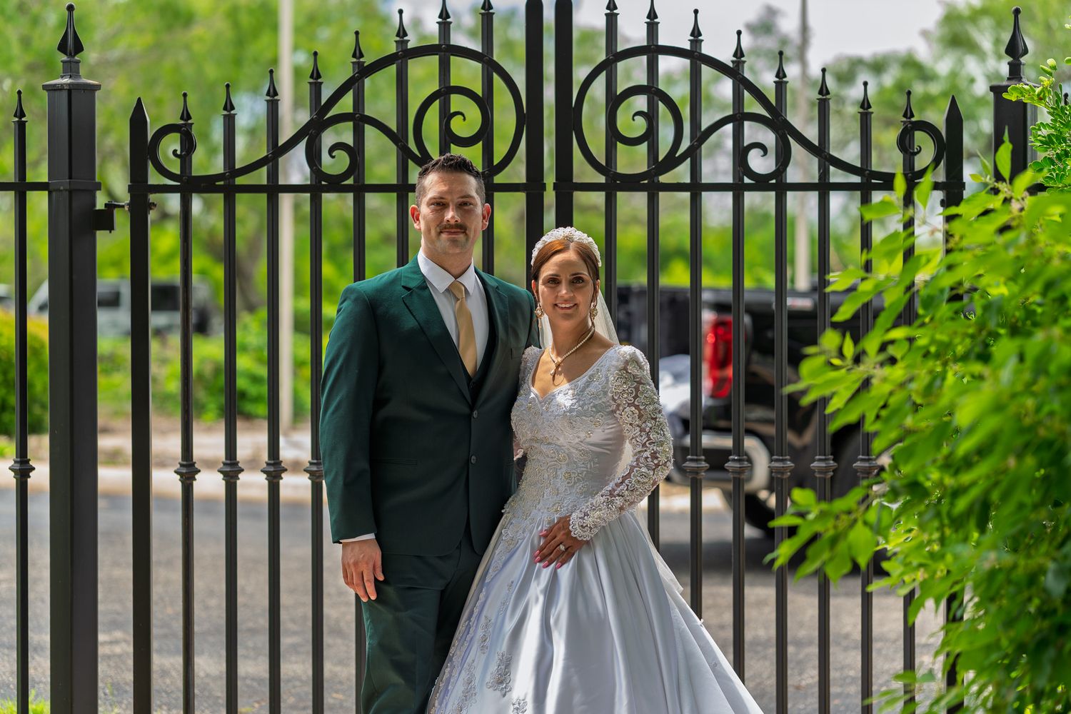 Bride and groom posing in front of ornate black iron gates, bride in white gown, groom in dark green suit.
