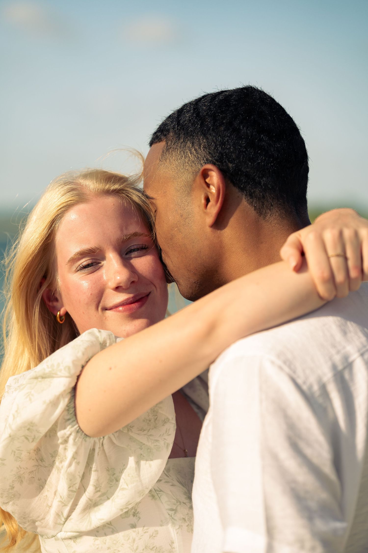 A young interracial couple embracing outdoors, blonde woman smiling as man kisses her cheek under a blue sky.
