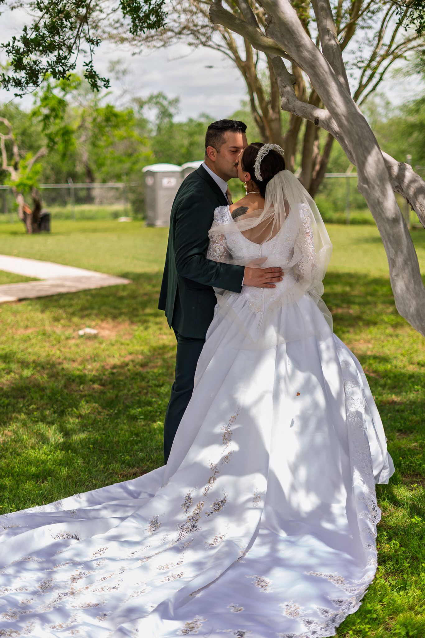 A bride and groom share a romantic kiss outdoors, with her elegant white gown and long floral train spread across green grass.