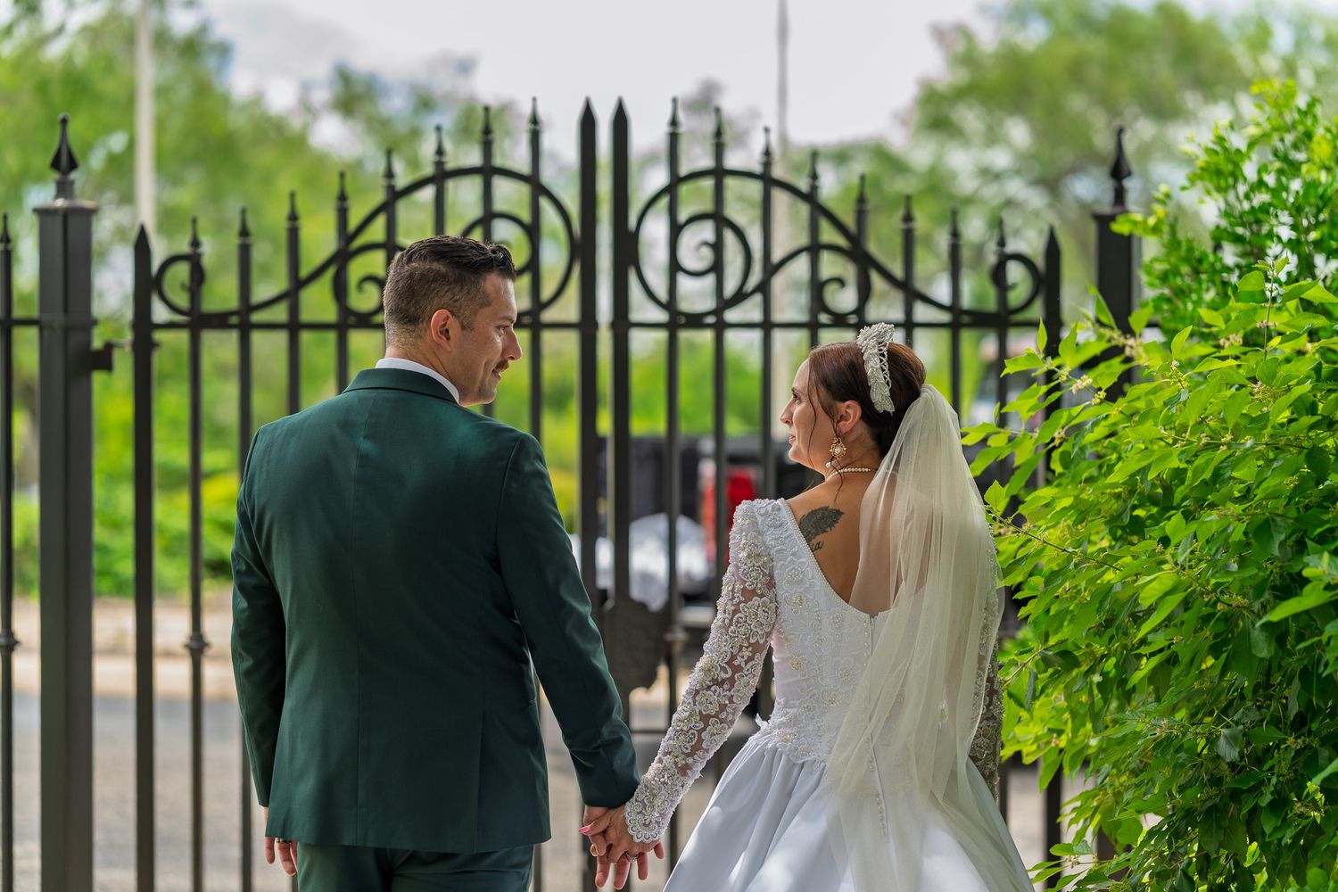 Bride and groom holding hands walking toward ornate iron gate surrounded by lush greenery on their wedding day.