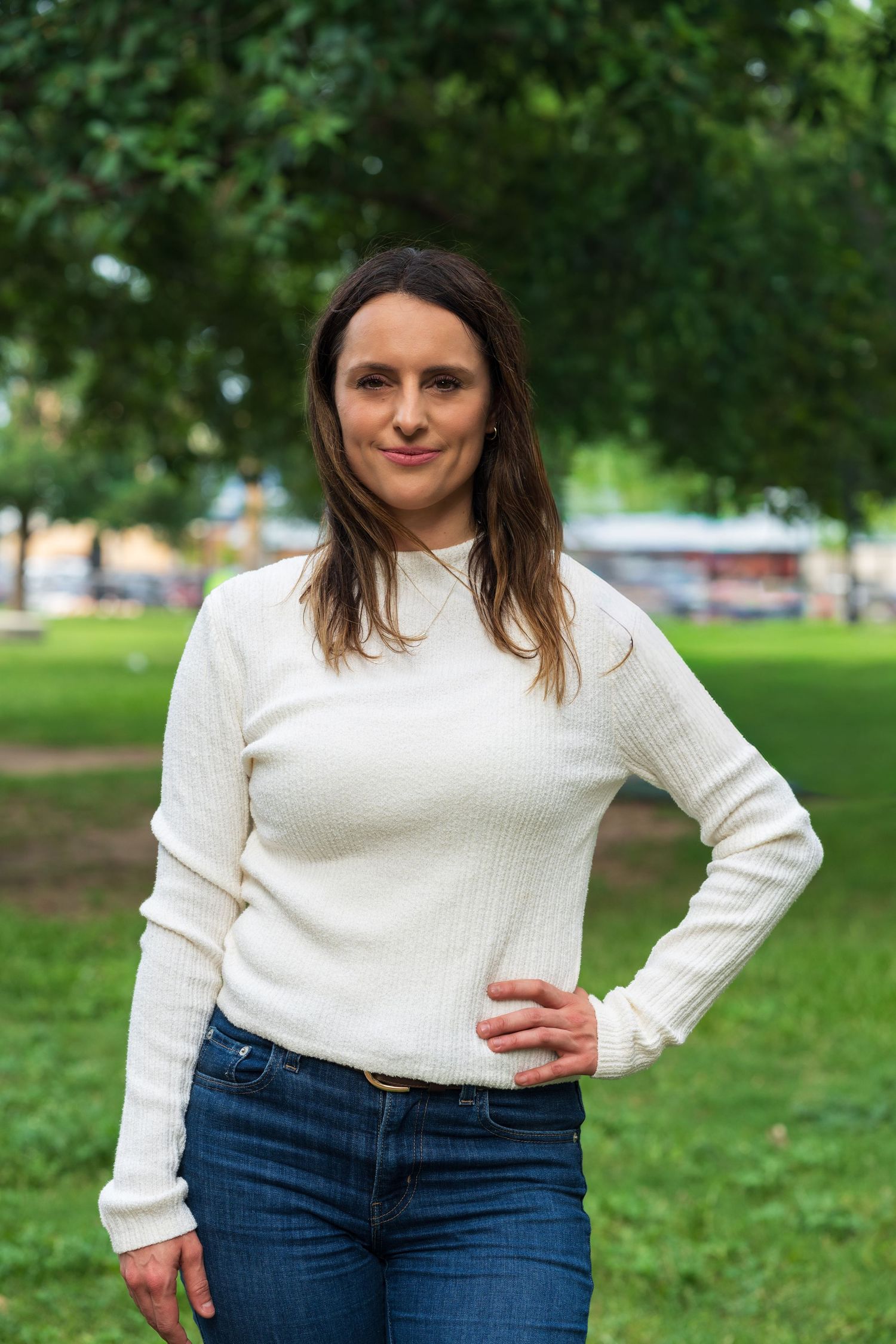 Woman in white sweater and jeans standing in a green park, smiling with hand on hip.