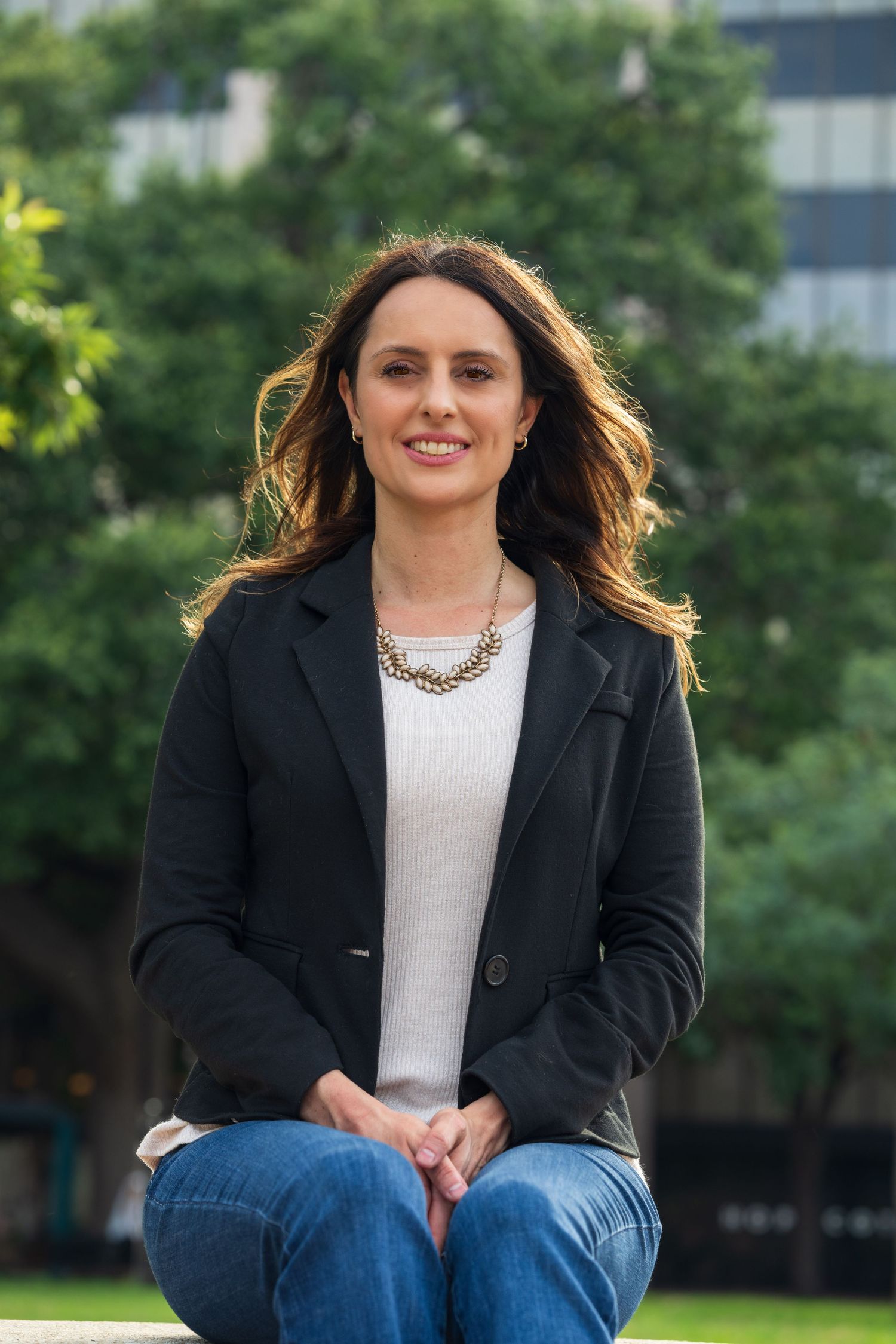 Woman in black blazer and jeans sitting outdoors in a park, smiling confidently.