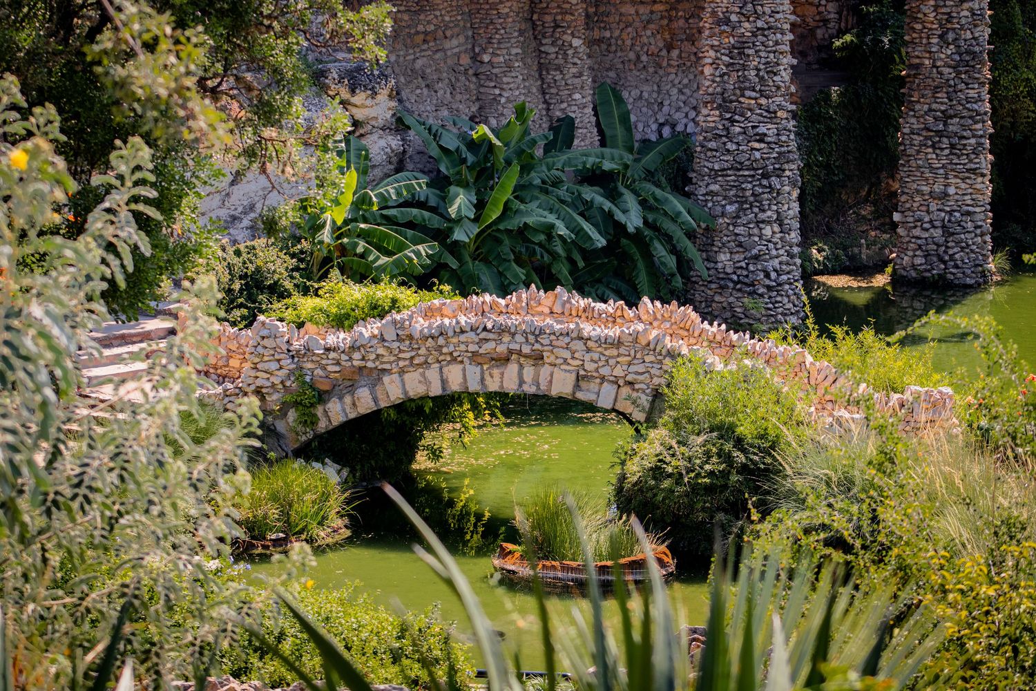 Stone arch bridge over a tranquil garden pond surrounded by lush tropical plants and ancient stone walls.