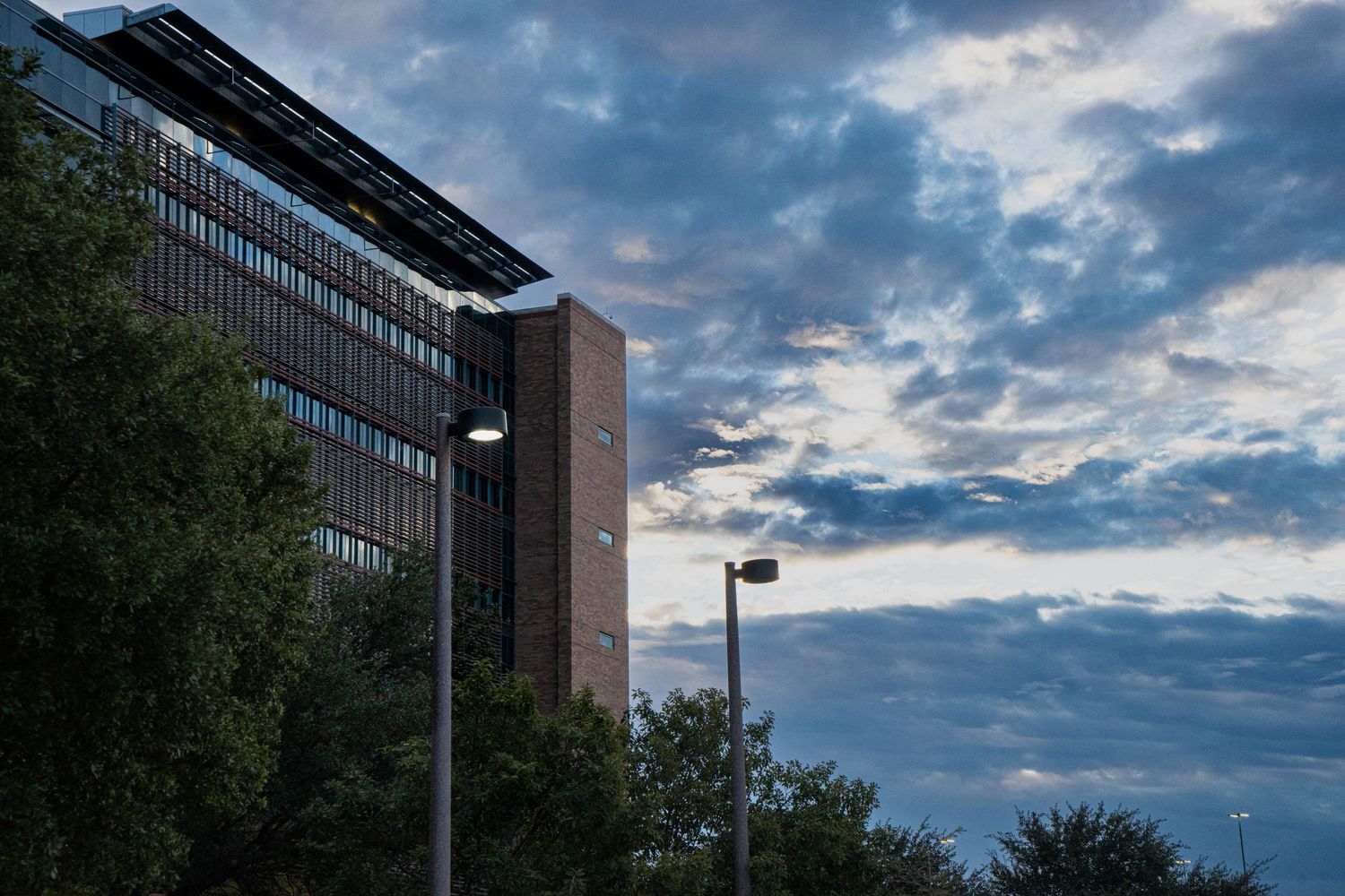 Tall brick office building with solar panels at dusk, framed by trees and street lights under dramatic cloudy sky.