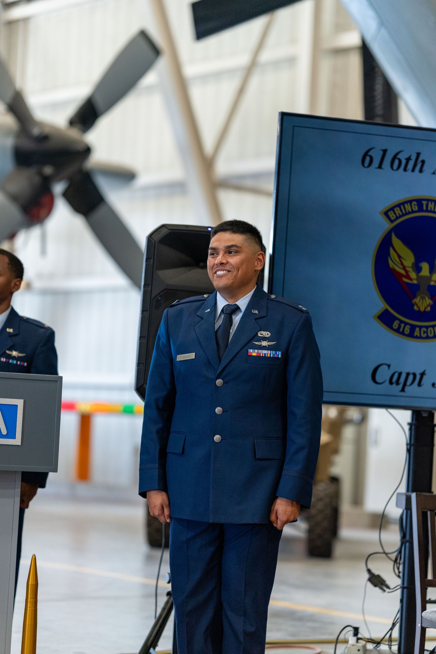 US Air Force officer in dress blues smiles at 616th AC2 Squadron ceremony in aircraft hangar.