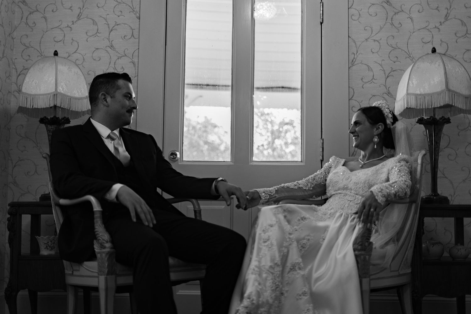 Black and white wedding photo of bride and groom holding hands while seated in elegant chairs by a window.