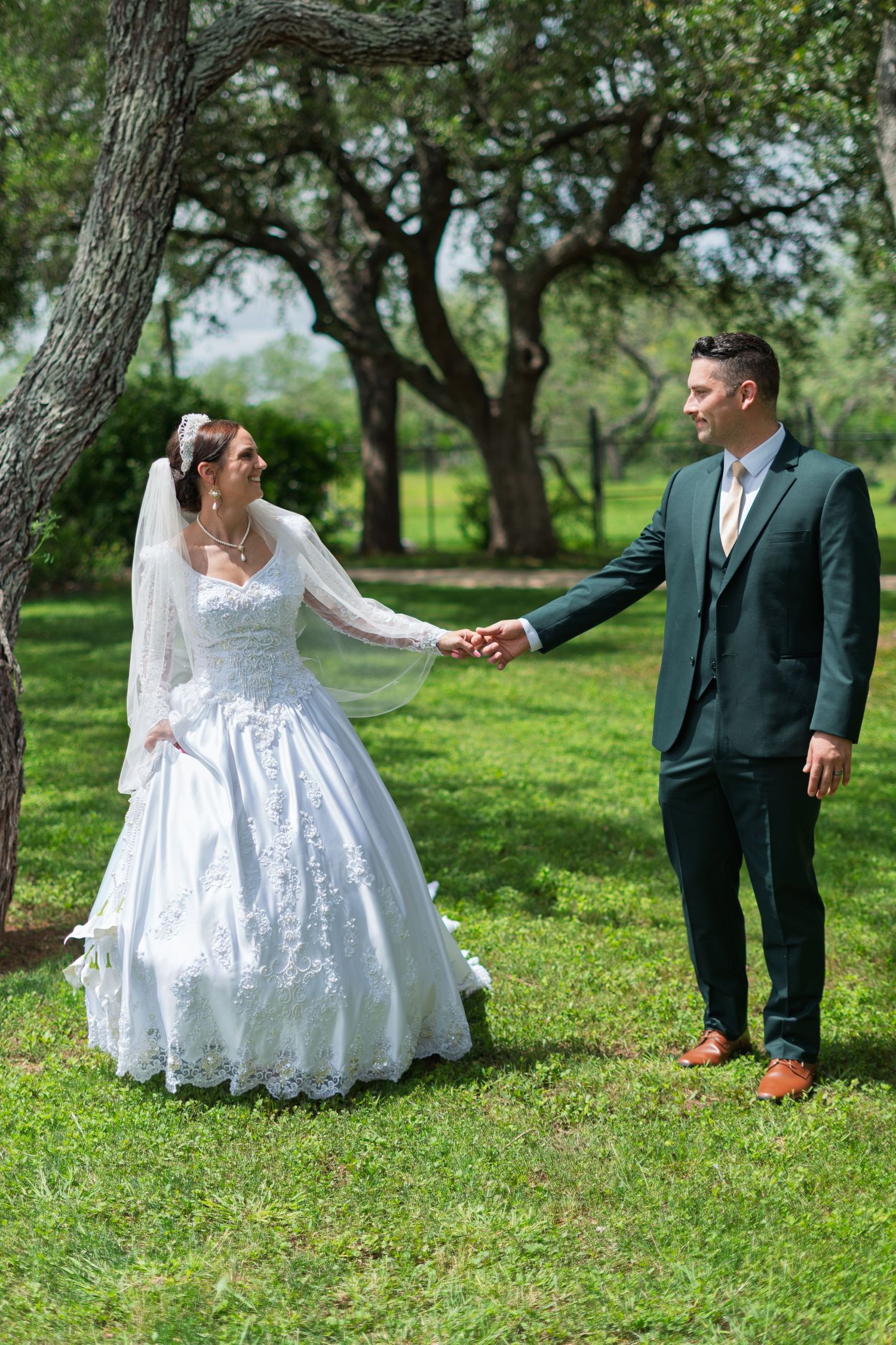 Bride in white lace gown and groom in dark green suit hold hands in a lush green park on their wedding day.