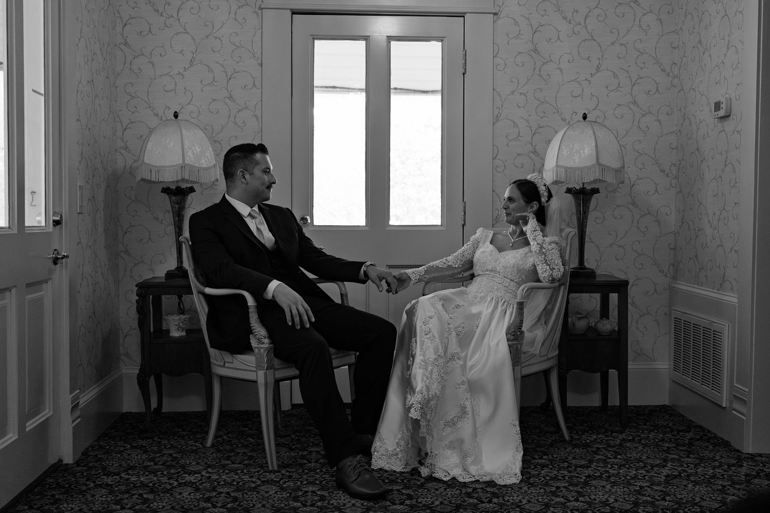 Black and white photo of bride and groom holding hands seated in elegant room with vintage lamps and window.