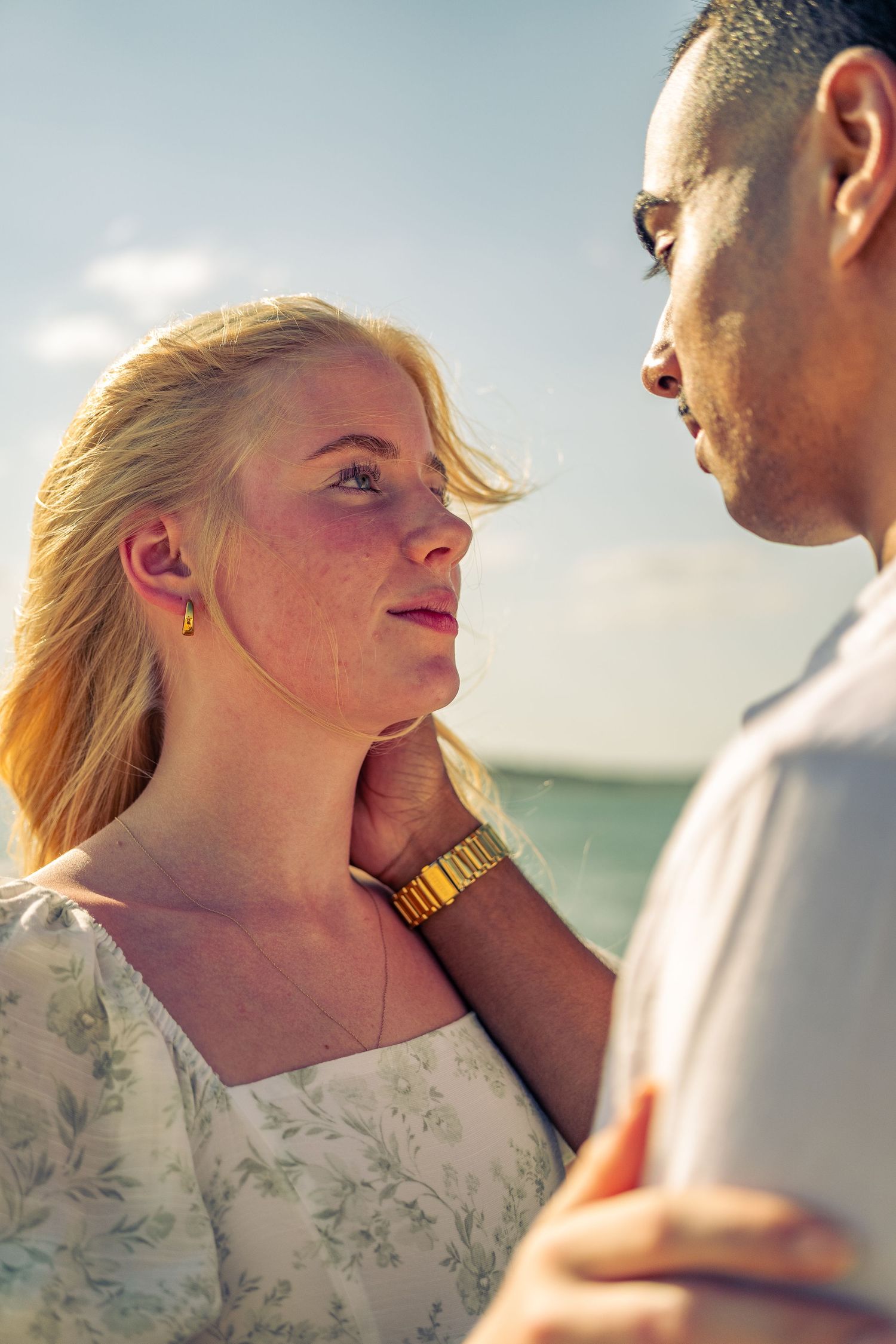 A couple shares a romantic moment by the ocean, with a blonde woman gazing up at a man resting his hand on her neck.