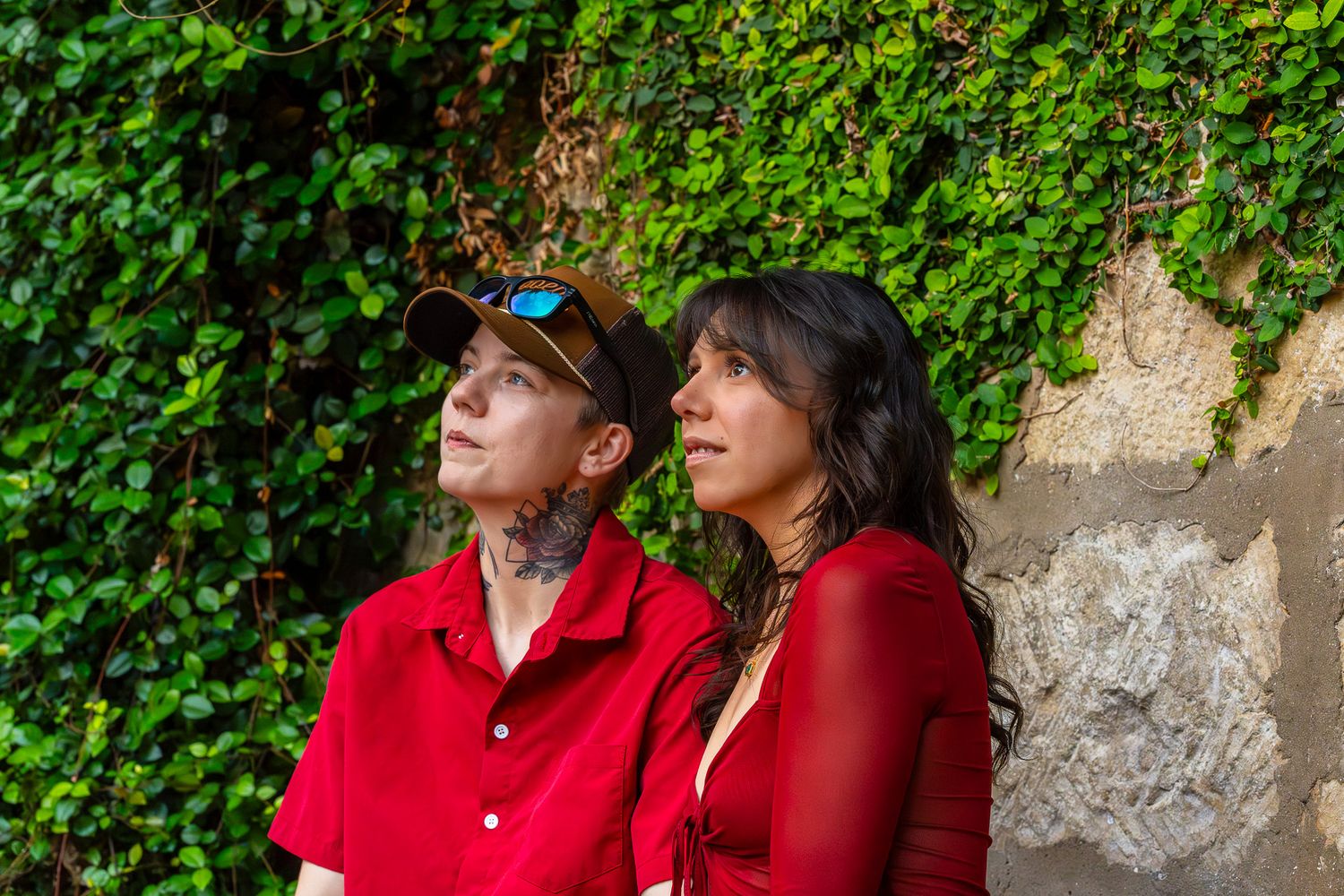 Couple in matching red tops gazing upward against a lush green ivy-covered stone wall outdoors.