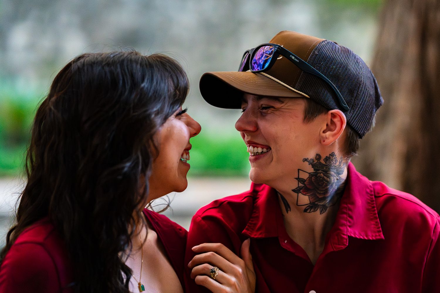 Two people with tattoos smiling at each other outdoors, wearing red shirts and a cap, sharing a joyful moment.