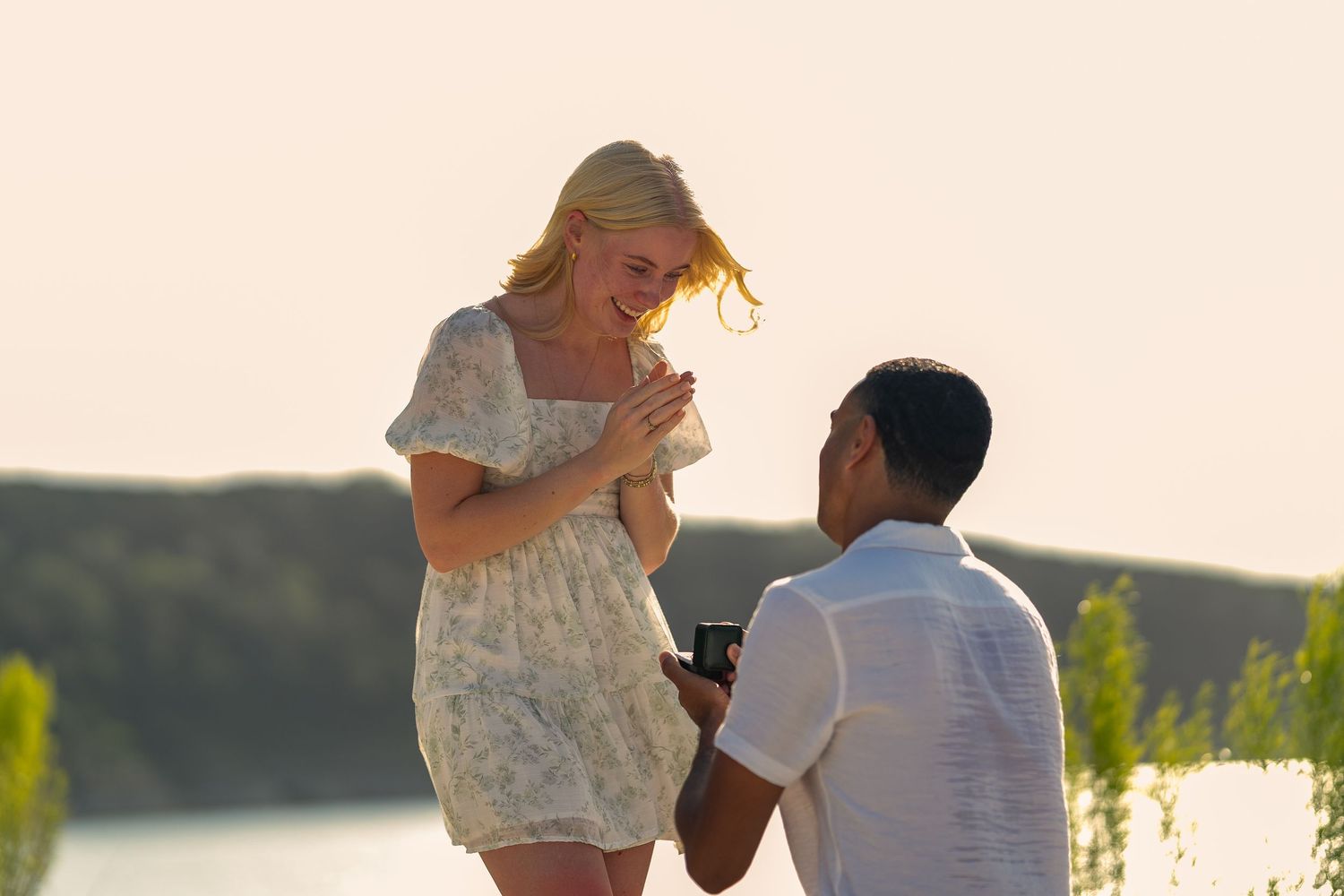Man kneeling to propose to a smiling blonde woman outdoors near a lake at sunset.