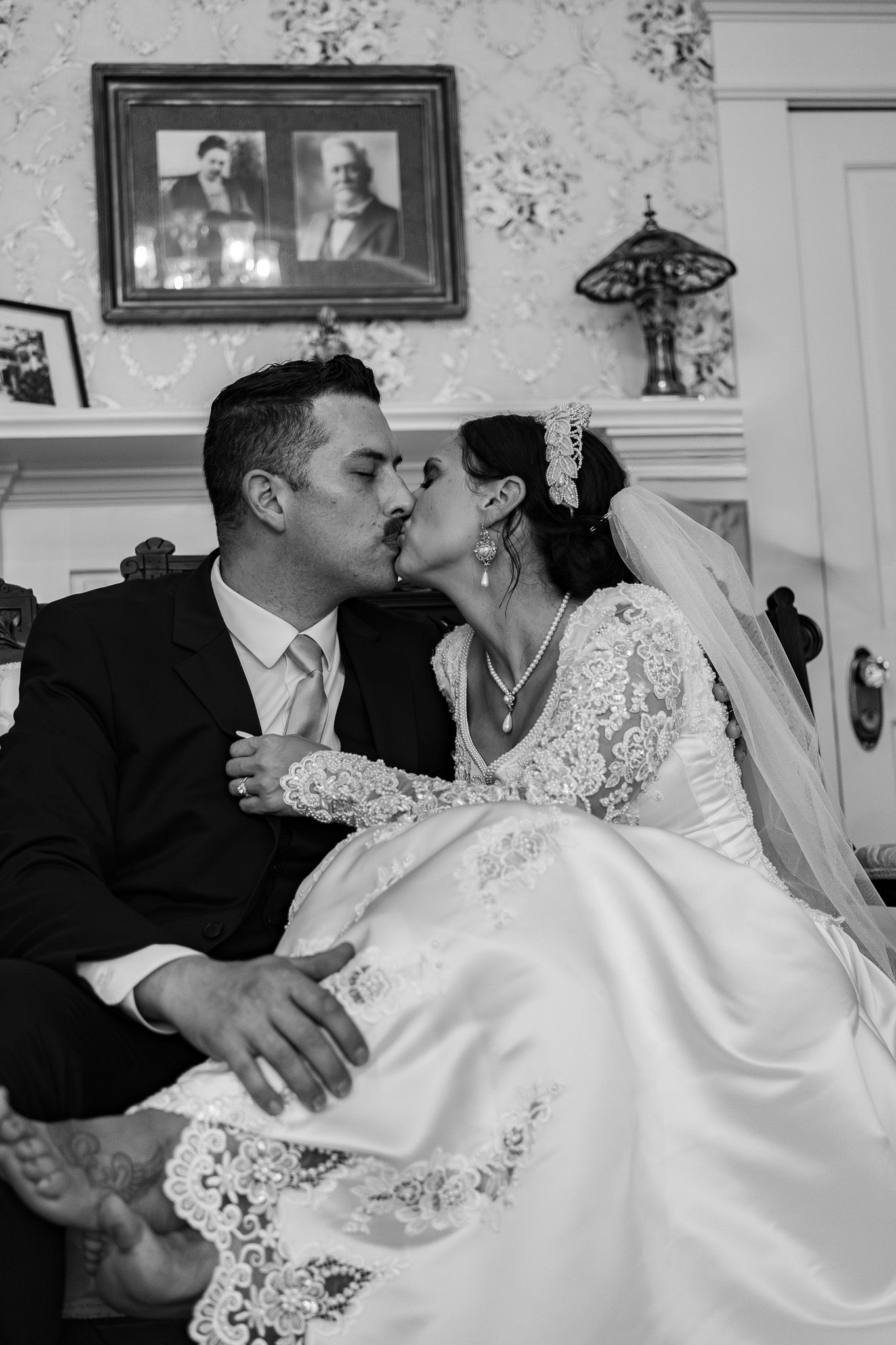 Black and white photo of bride and groom sharing a kiss while seated in a vintage decorated room.