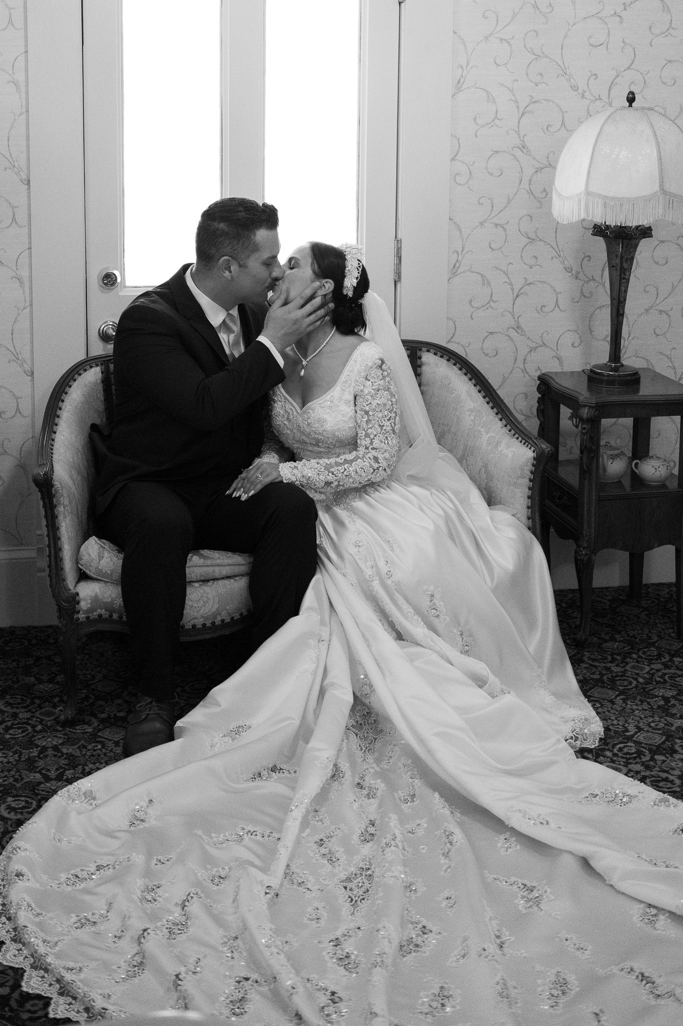 Black and white photo of a bride and groom sharing a romantic kiss while seated on an elegant vintage sofa.