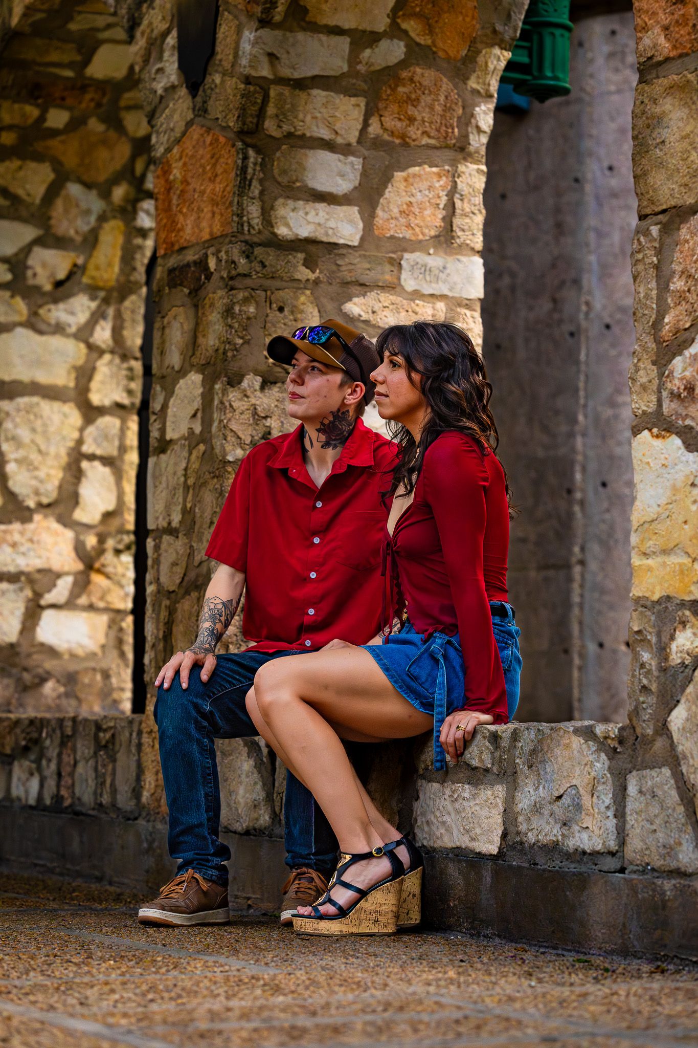 Couple in coordinating red outfits sitting together against a rustic stone wall architecture.