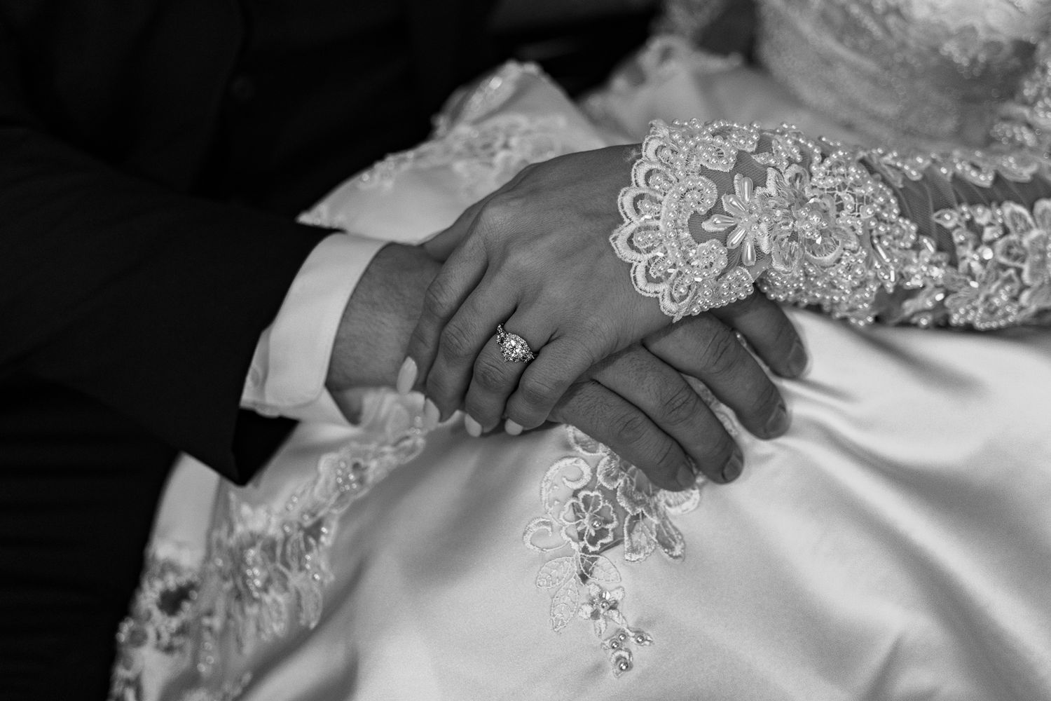 Black and white close-up of bride and groom holding hands showing wedding ring and lace dress detail.