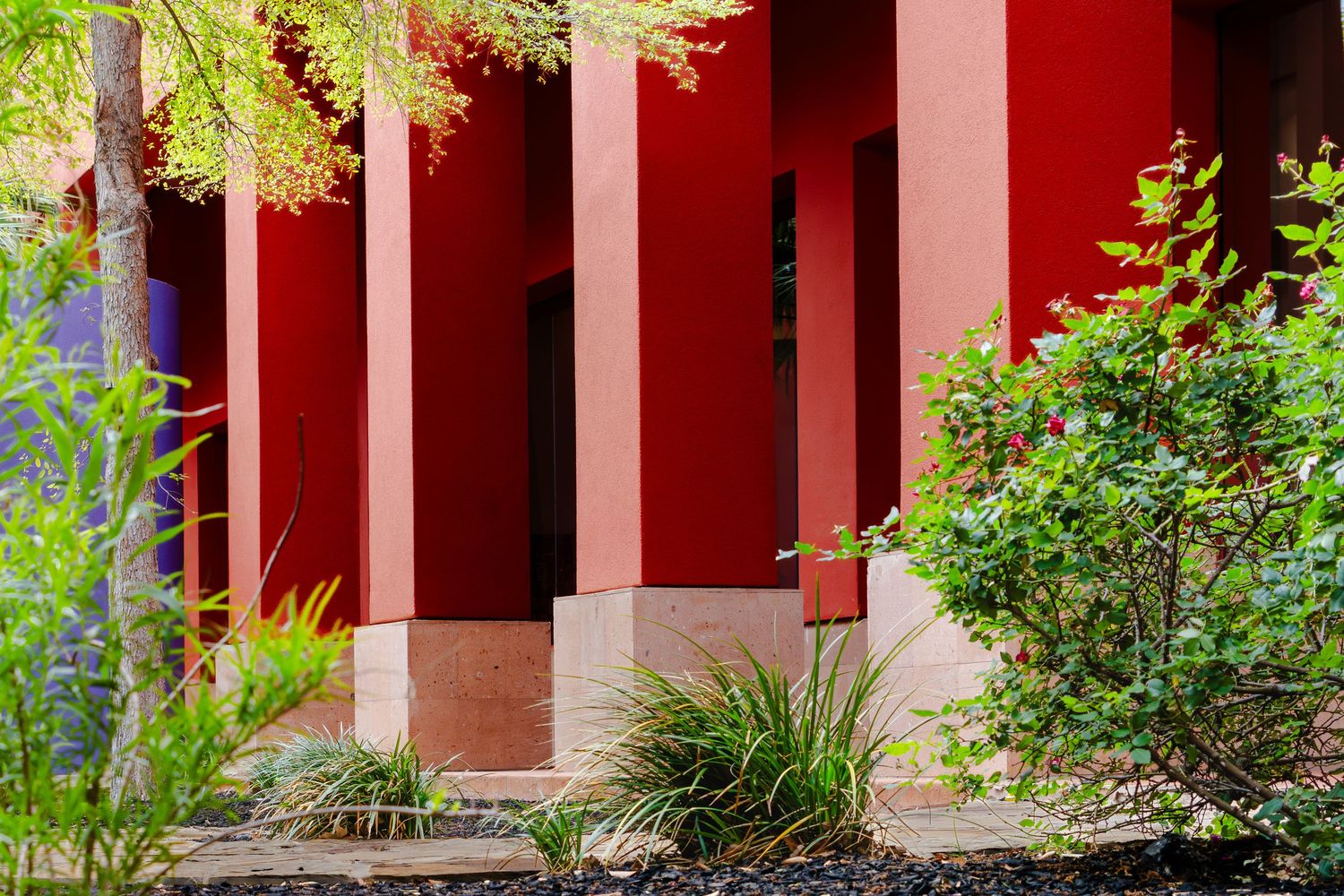 Red building exterior with tall columns surrounded by lush green shrubs and ornamental grasses in a landscaped garden.