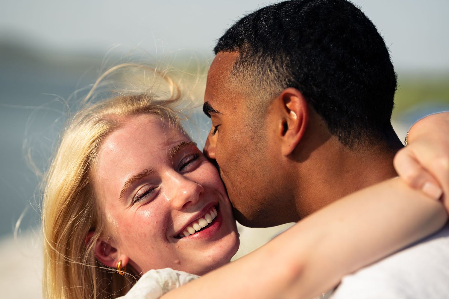 A happy interracial couple sharing a romantic kiss on a sunny beach, with the woman smiling joyfully.