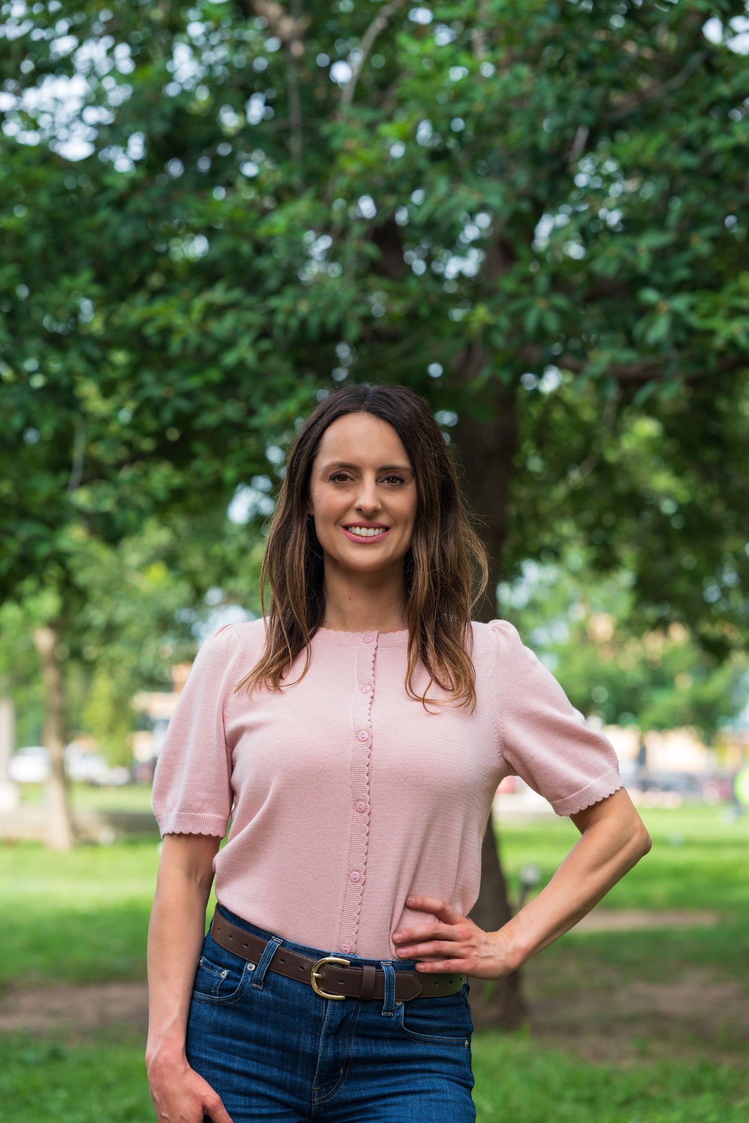 Woman in pink blouse and jeans standing in a park with hand on hip, smiling.
