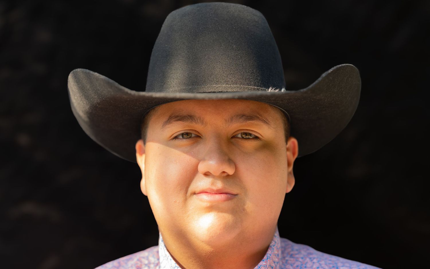 Man wearing a black cowboy hat and floral shirt smiling confidently against a dark background.