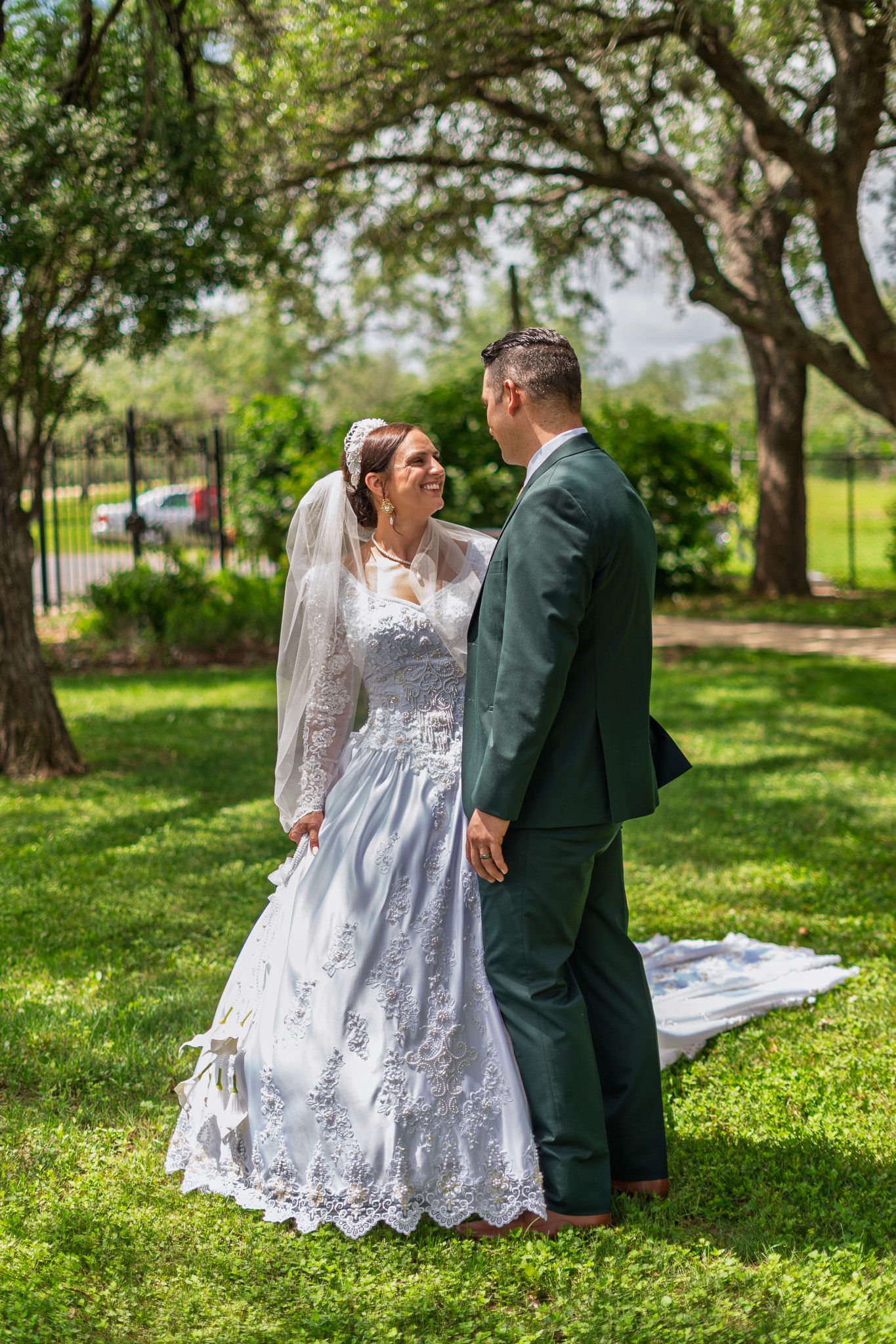 Bride and groom holding hands in lush green park, bride in lace wedding gown with veil, groom in dark suit.
