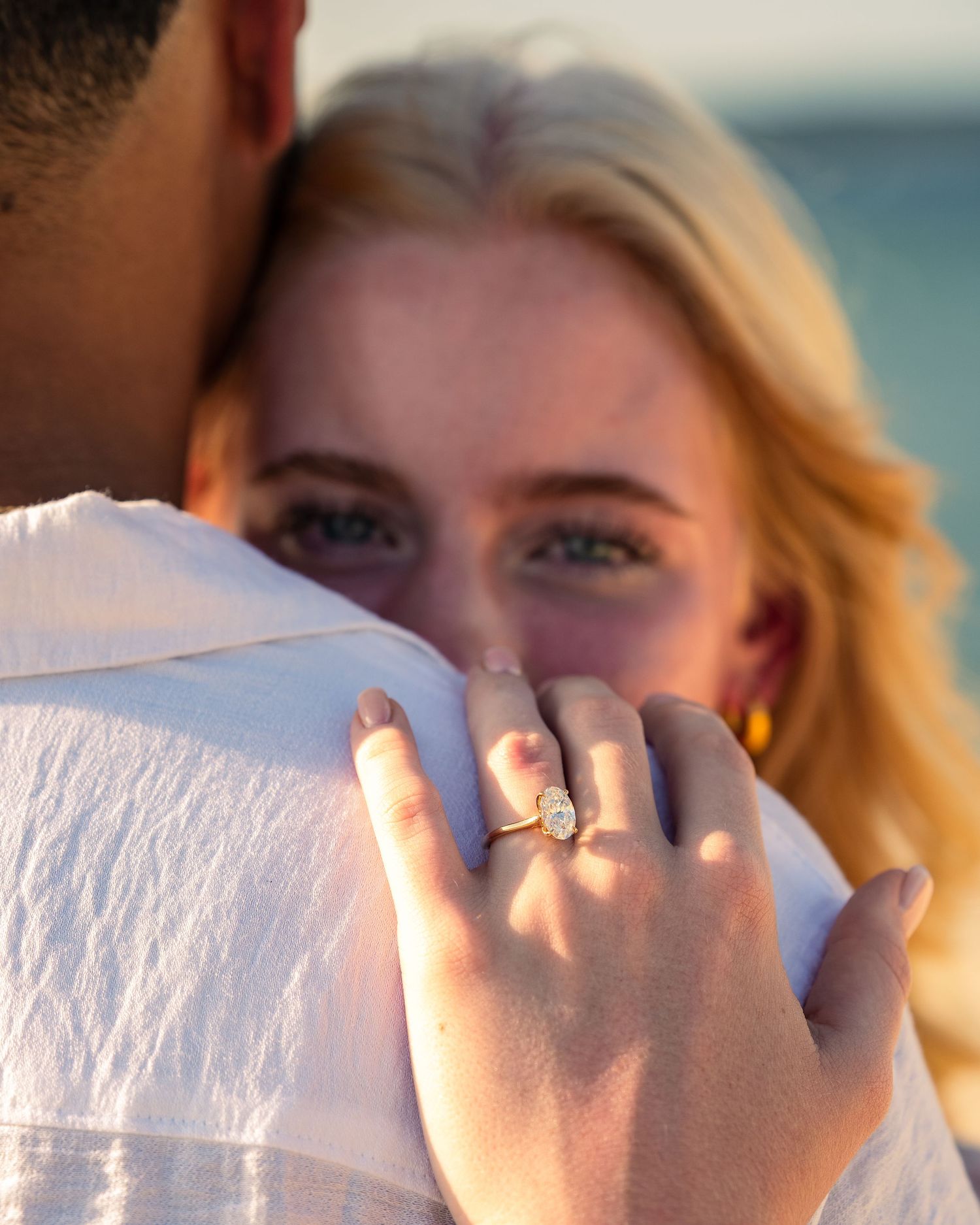 Blonde woman with blue eyes hugging a man, showing off a diamond engagement ring on her finger near the ocean.
