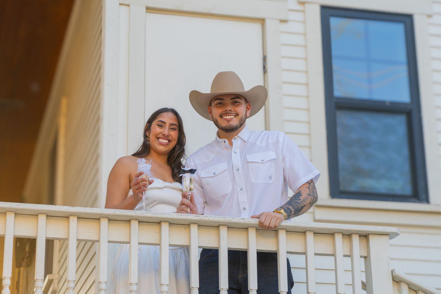 Smiling couple standing on a porch balcony, man wearing cowboy hat and tattoos, woman in white dress.