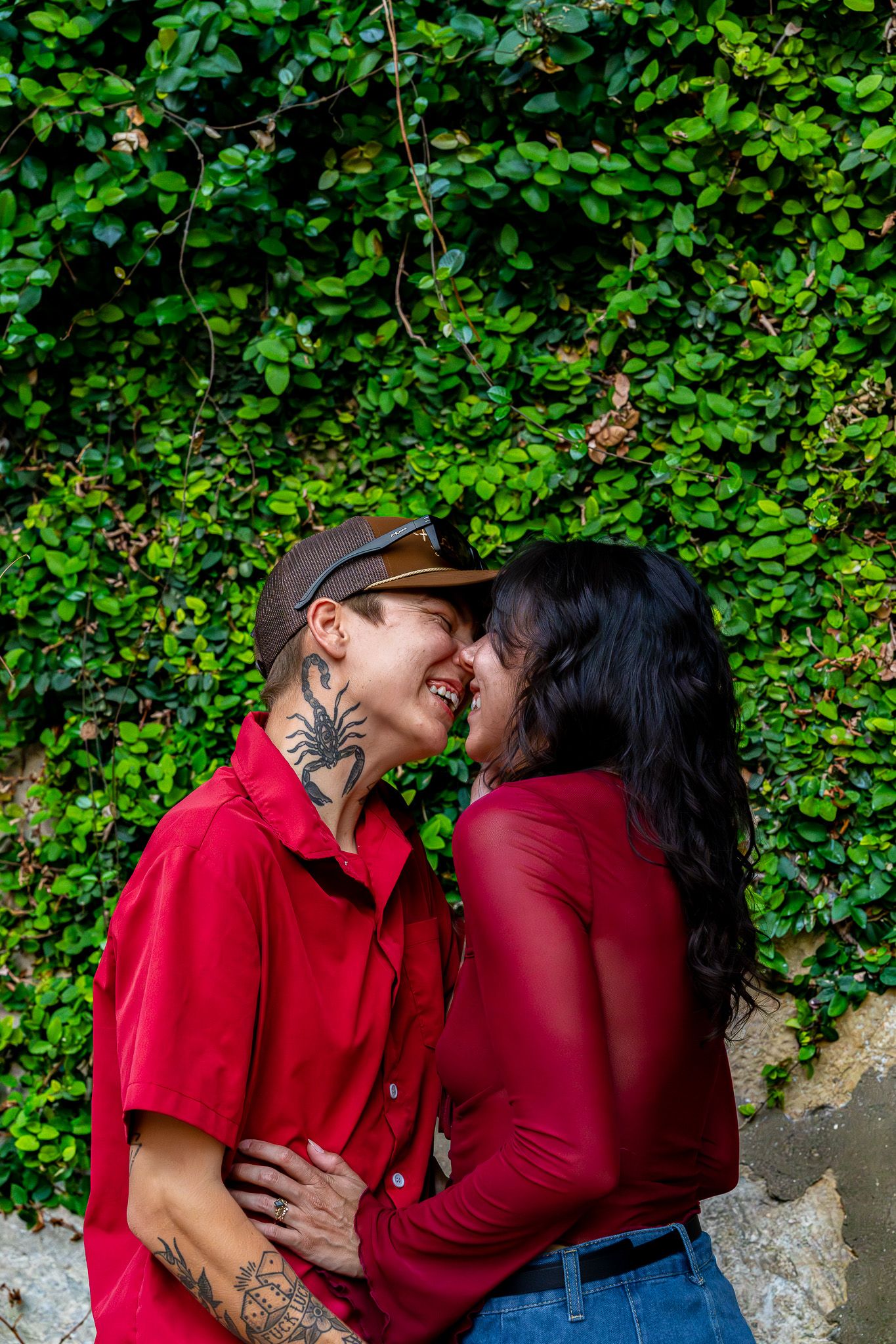 Two women in matching red shirts share a romantic moment in front of a lush green ivy wall.