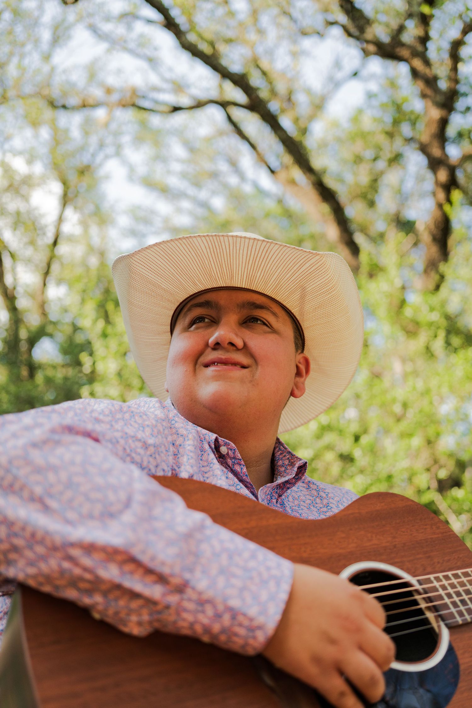 Young man in white cowboy hat playing acoustic guitar outdoors among green trees, wearing floral shirt.