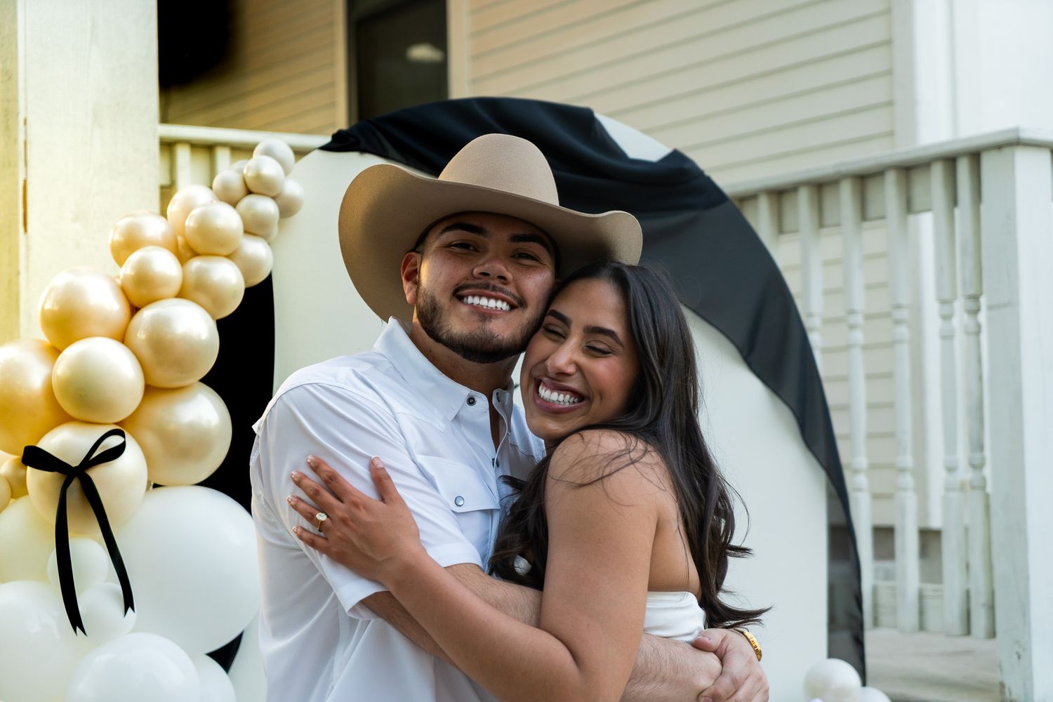 Couple smiling and hugging under umbrella near gold and black balloons during outdoor celebration.