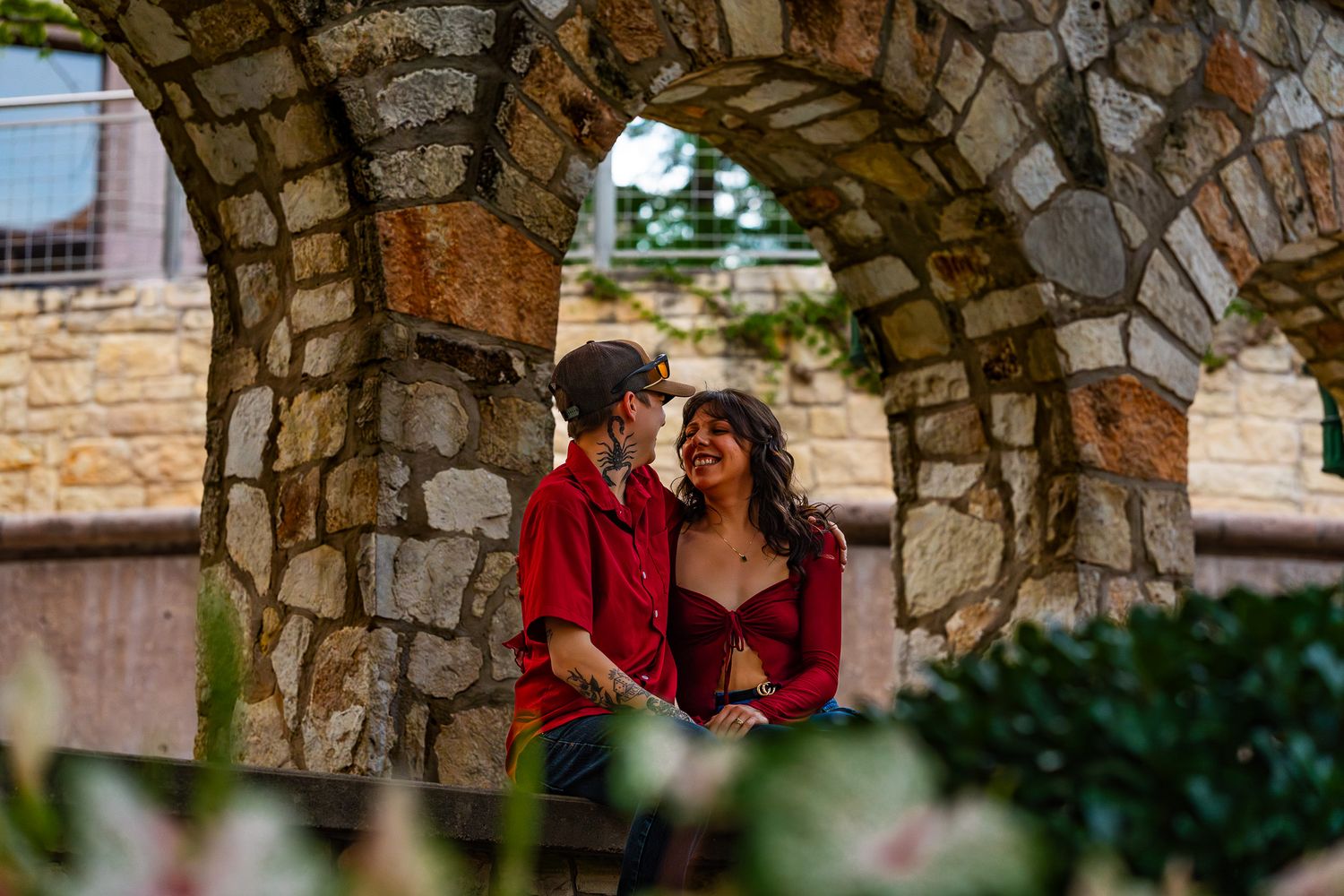 Couple in matching red outfits sharing a romantic moment under a stone arch in a garden setting.
