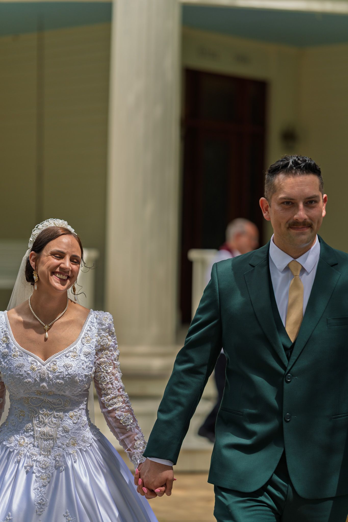 Smiling bride in white lace gown and groom in green suit holding hands outside a church after wedding ceremony.