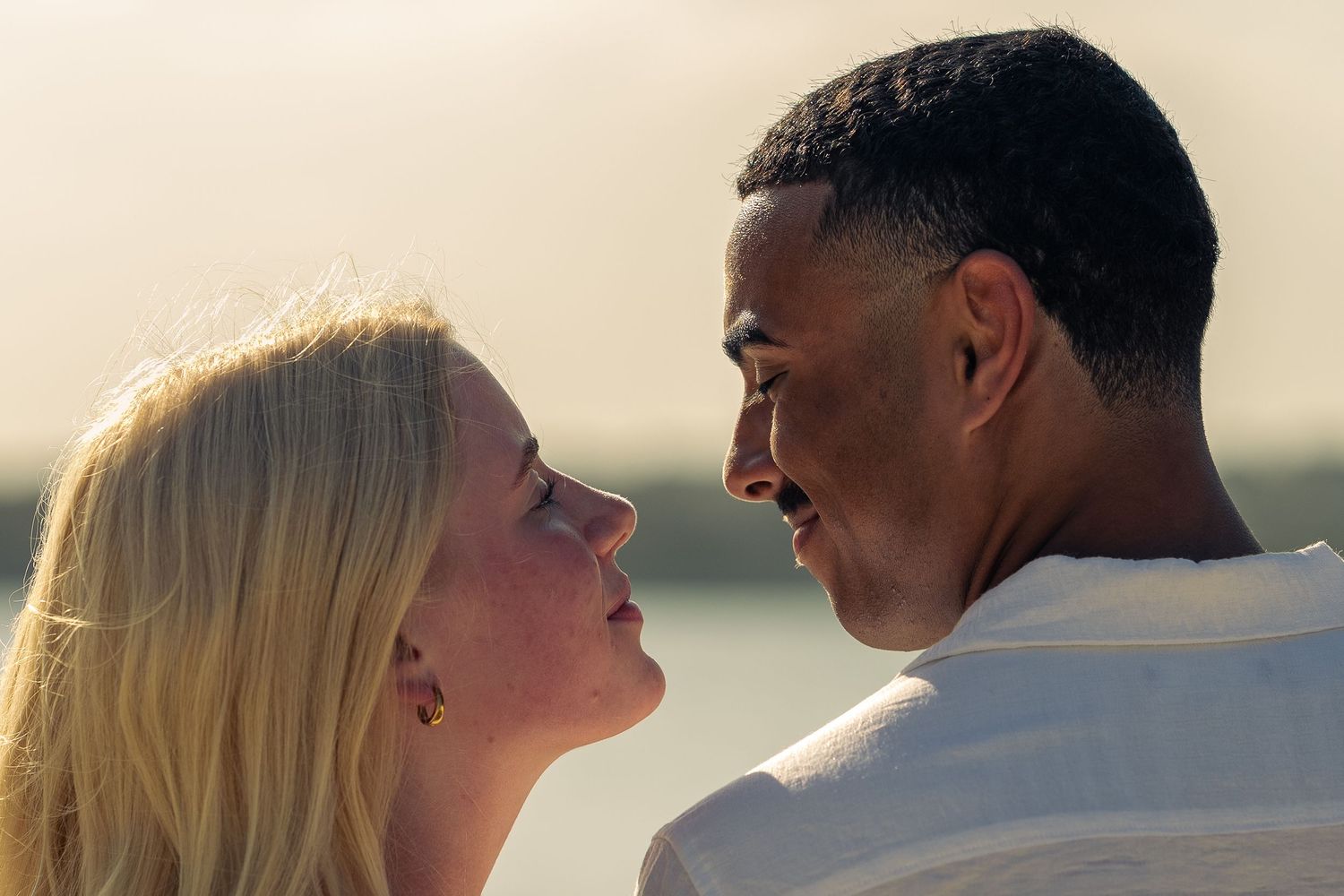A blonde woman and a man share an intimate moment outdoors near water in warm golden light.
