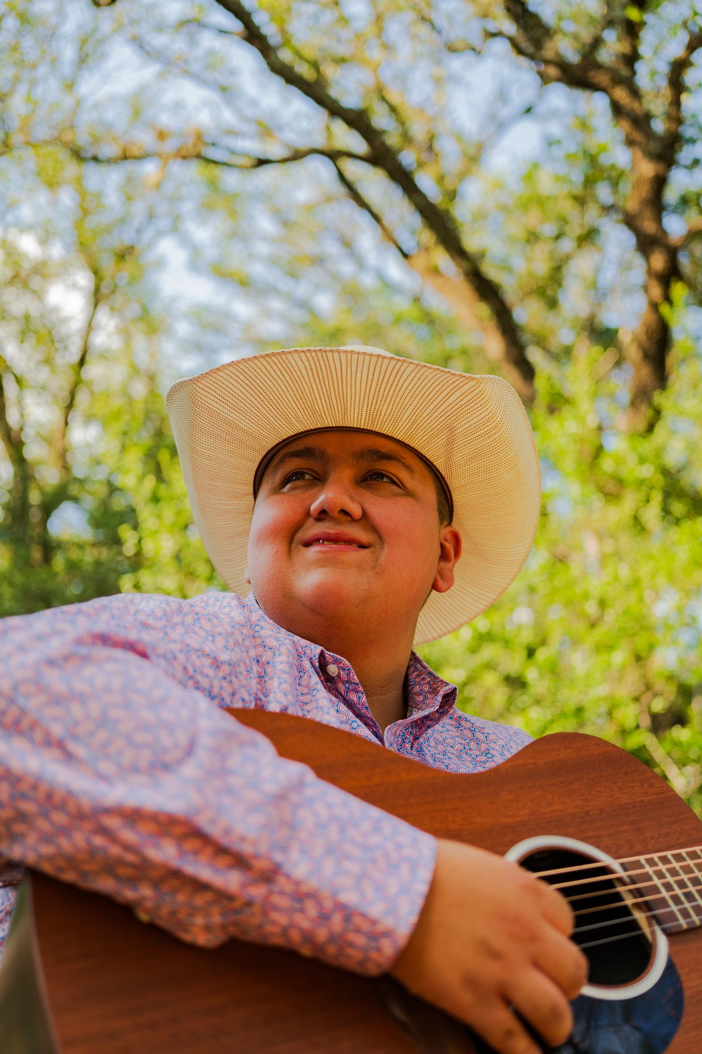 Young man in cowboy hat playing acoustic guitar outdoors, smiling upward surrounded by lush green trees.
