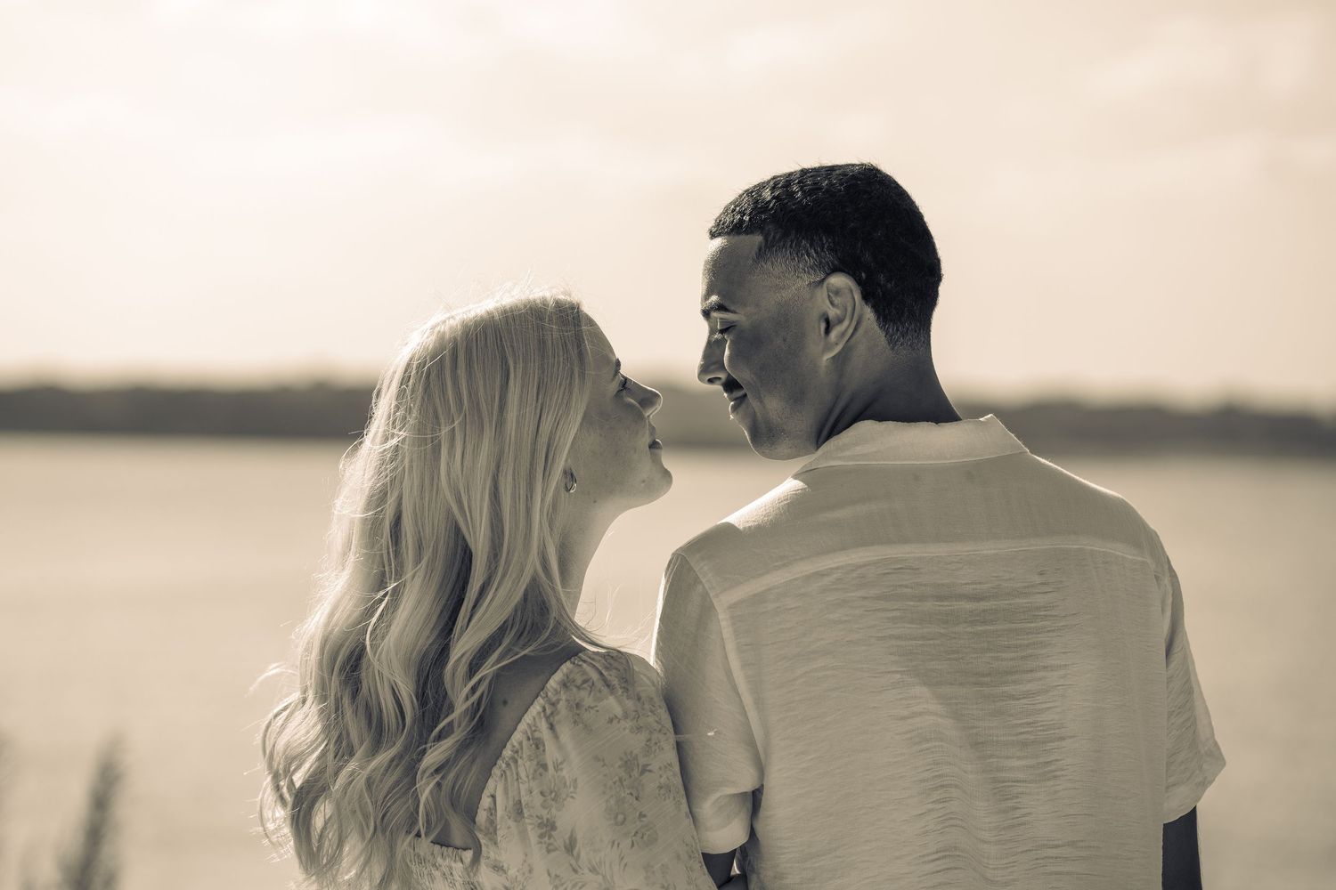 Romantic couple sharing an intimate moment near a serene lake at sunset in a soft sepia-toned photo.