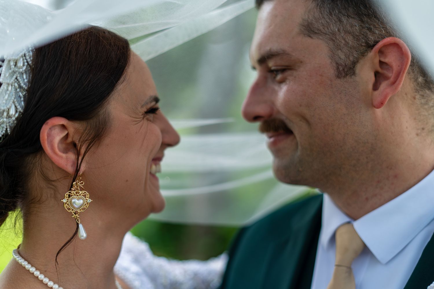 Close-up of smiling bride and groom looking at each other under flowing wedding veil outdoors.