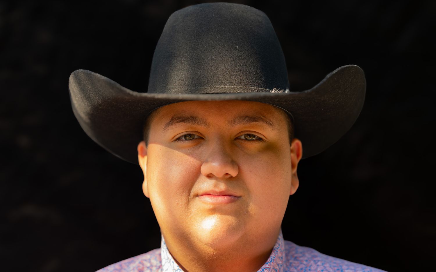 Man wearing a black cowboy hat and floral shirt posing against a dark background.