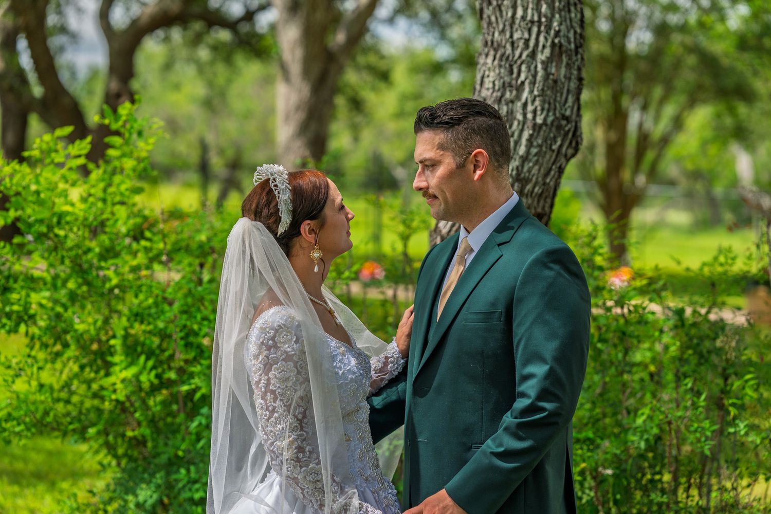 Bride and groom share a romantic moment outdoors, surrounded by lush green trees, bride in lace gown with veil.