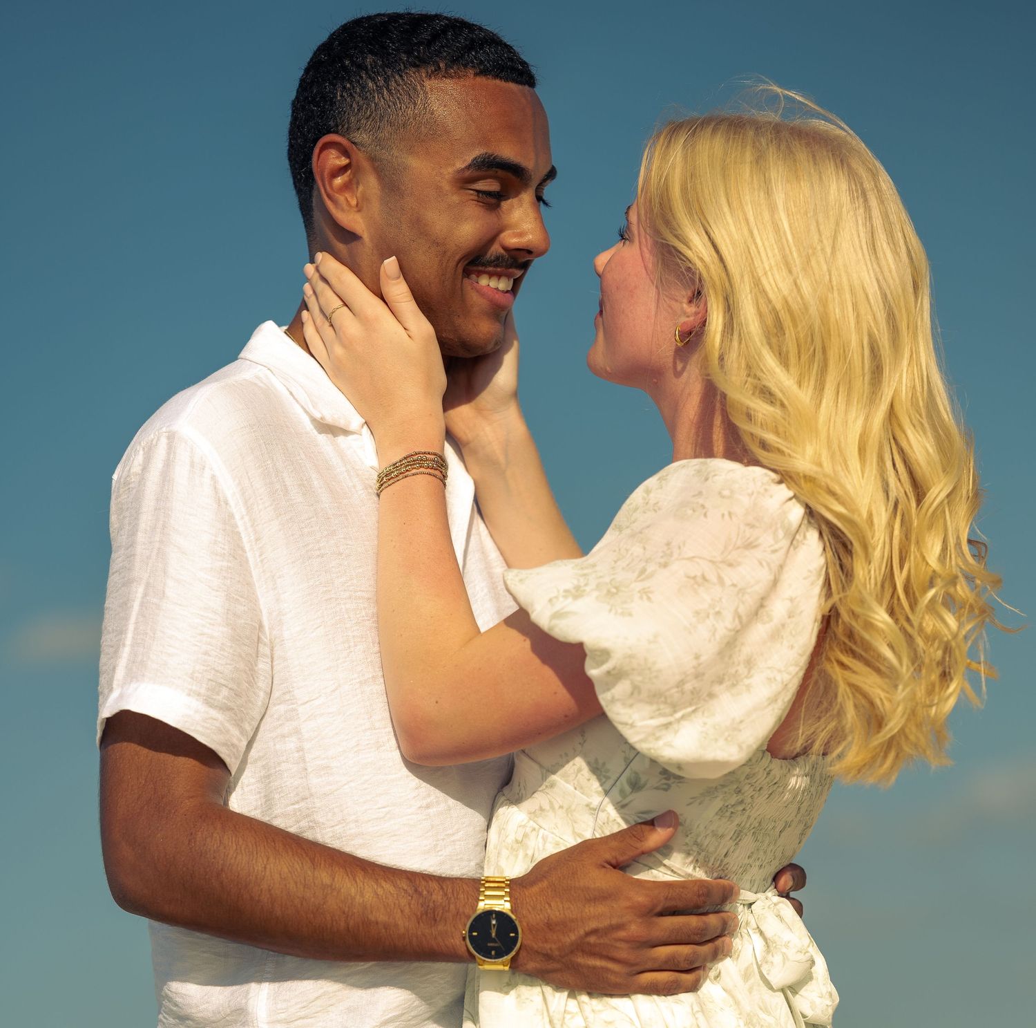 A joyful interracial couple embraces and smiles at each other against a blue sky backdrop.