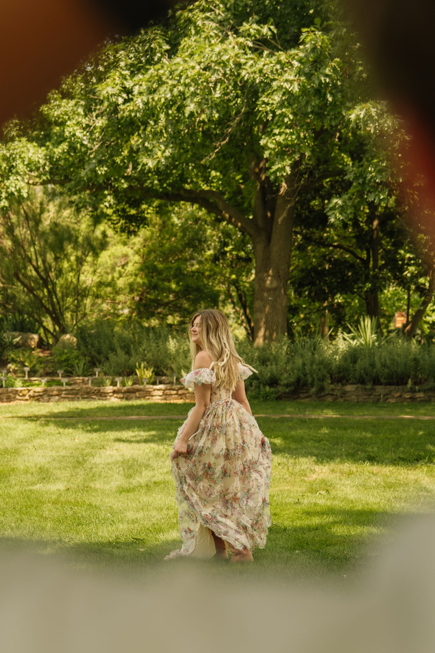 Woman in floral off-shoulder maxi dress twirling in lush green garden with large trees.
