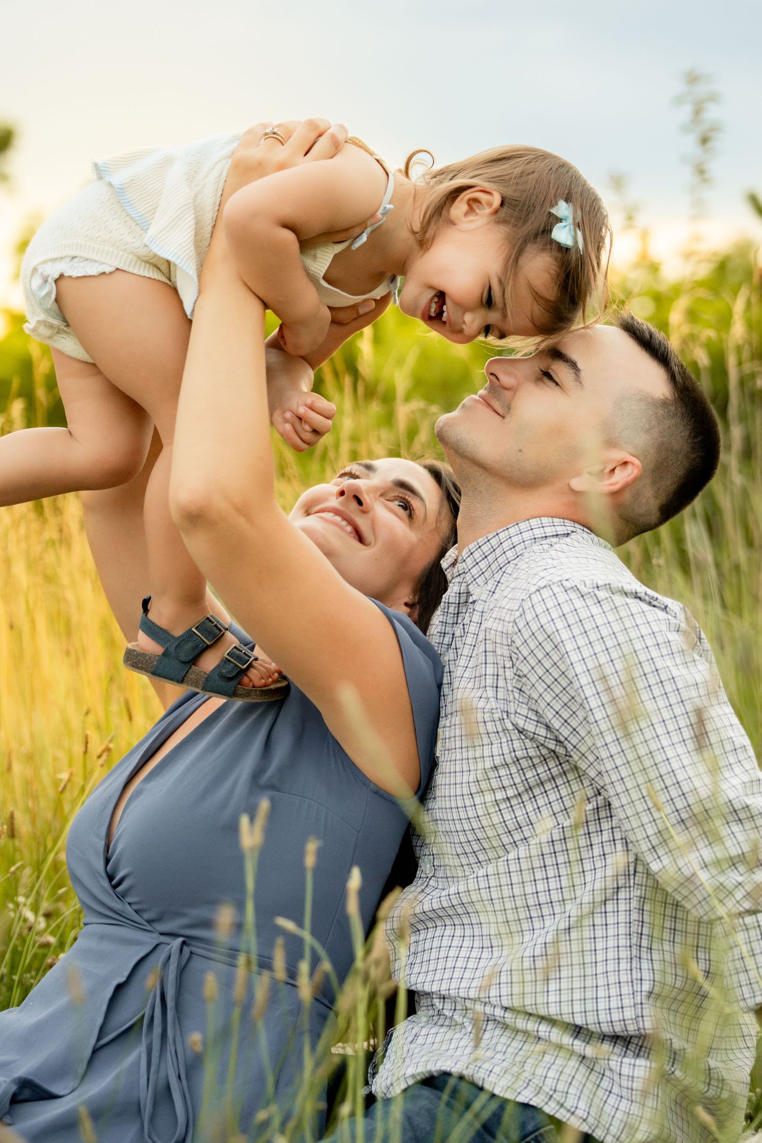 Happy family with parents lifting their toddler daughter in a sunny green field, sharing joyful smiles outdoors.
