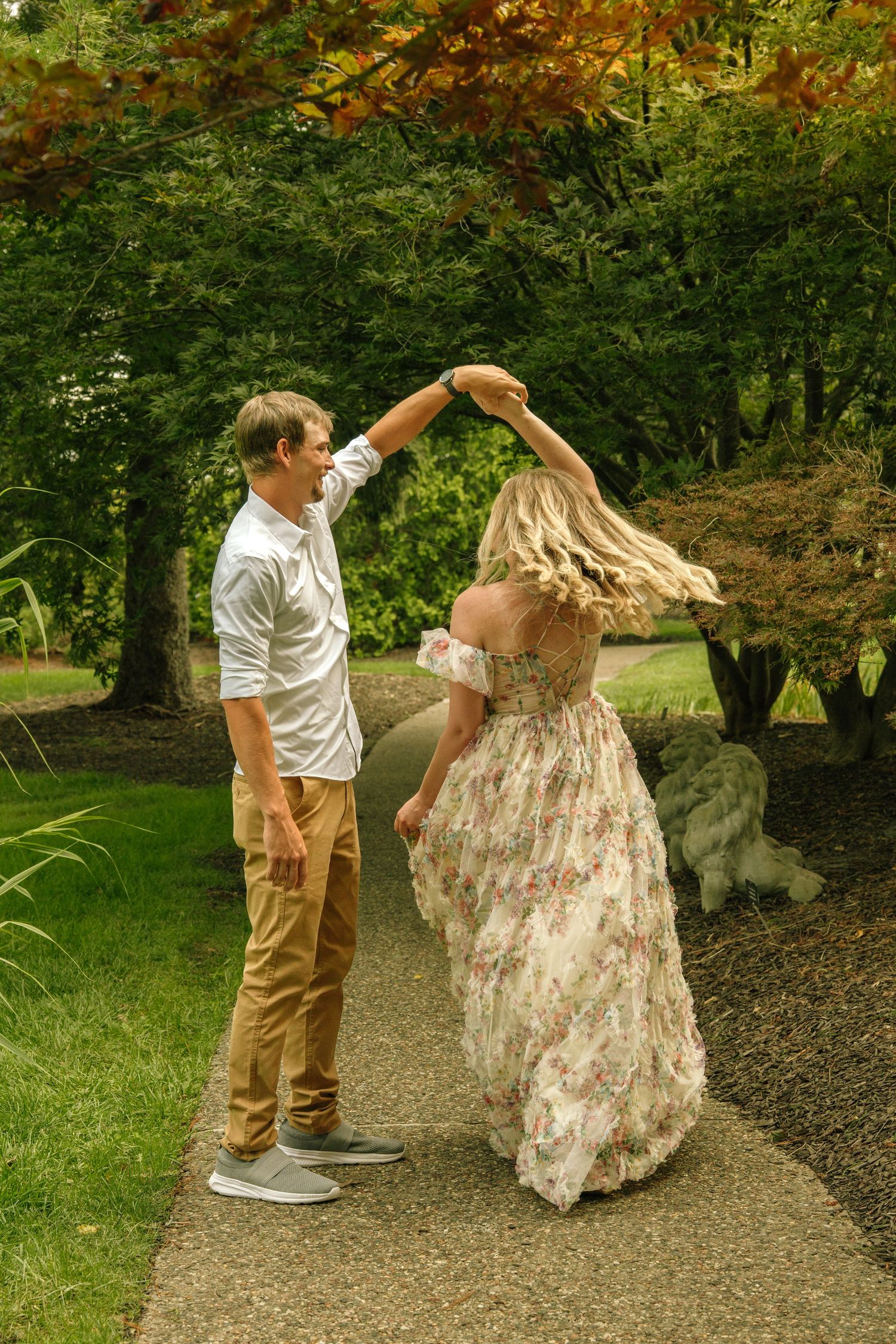 Man spinning woman in a floral maxi dress on a garden path surrounded by colorful autumn trees.