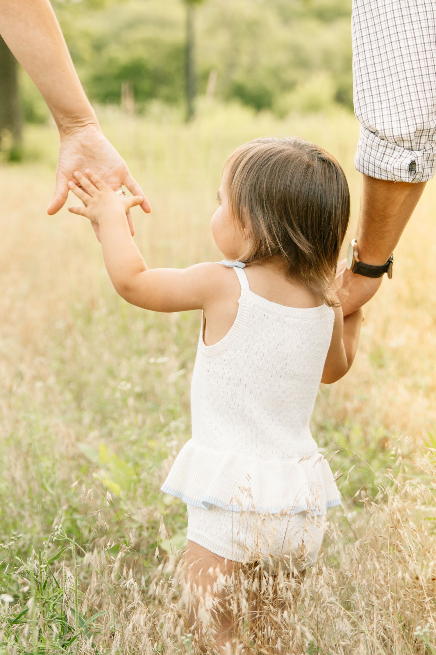 A toddler in a white dress walks through a sunny field, holding hands with two adults on either side.